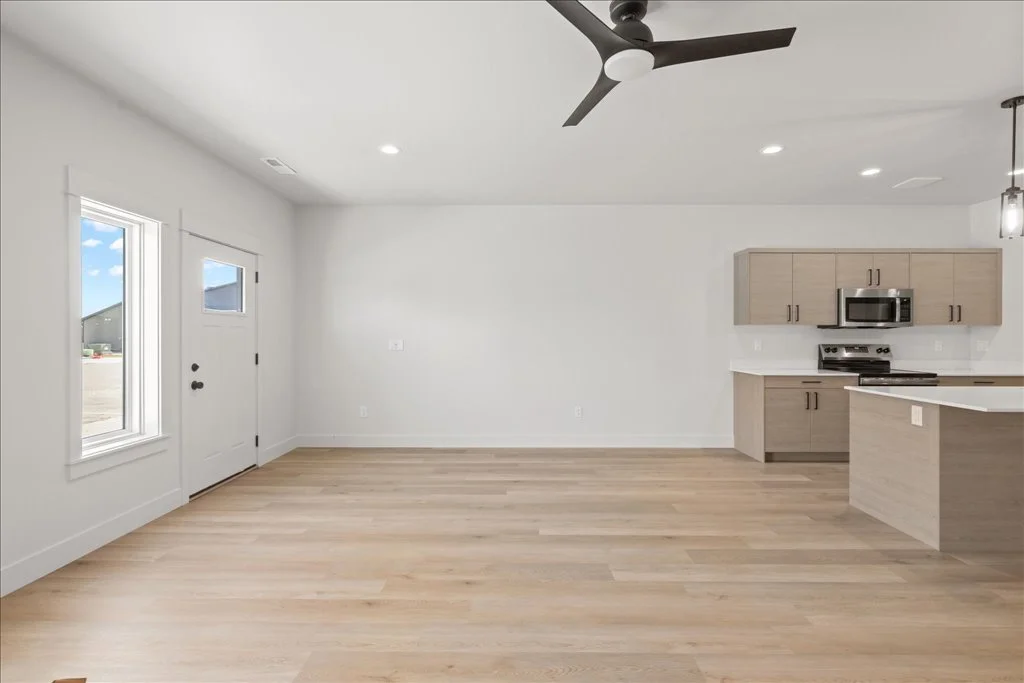 Empty living room and kitchen with light wood flooring, white walls, a ceiling fan, a window, a door, and kitchen cabinets with stainless steel appliances.