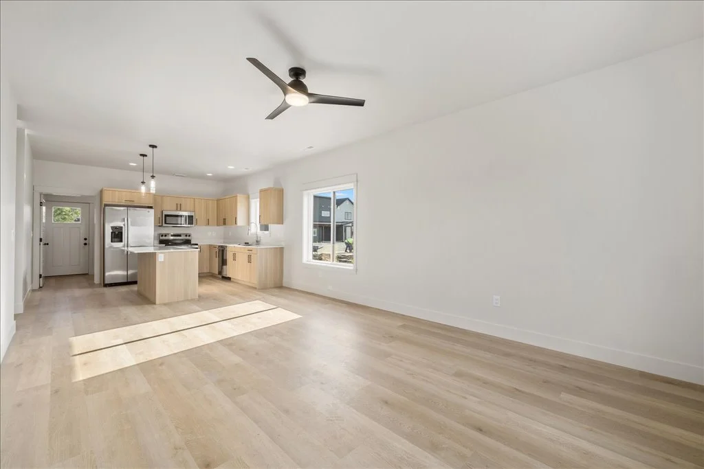 Empty living room with light wood flooring, white walls, a ceiling fan, and a view of the kitchen with light wood cabinets, stainless steel appliances, and a large window.