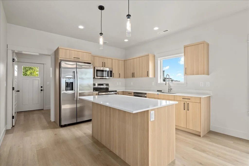 Modern kitchen with light wood cabinets, stainless steel appliances, and a white countertop island, with a window overlooking blue sky and trees.