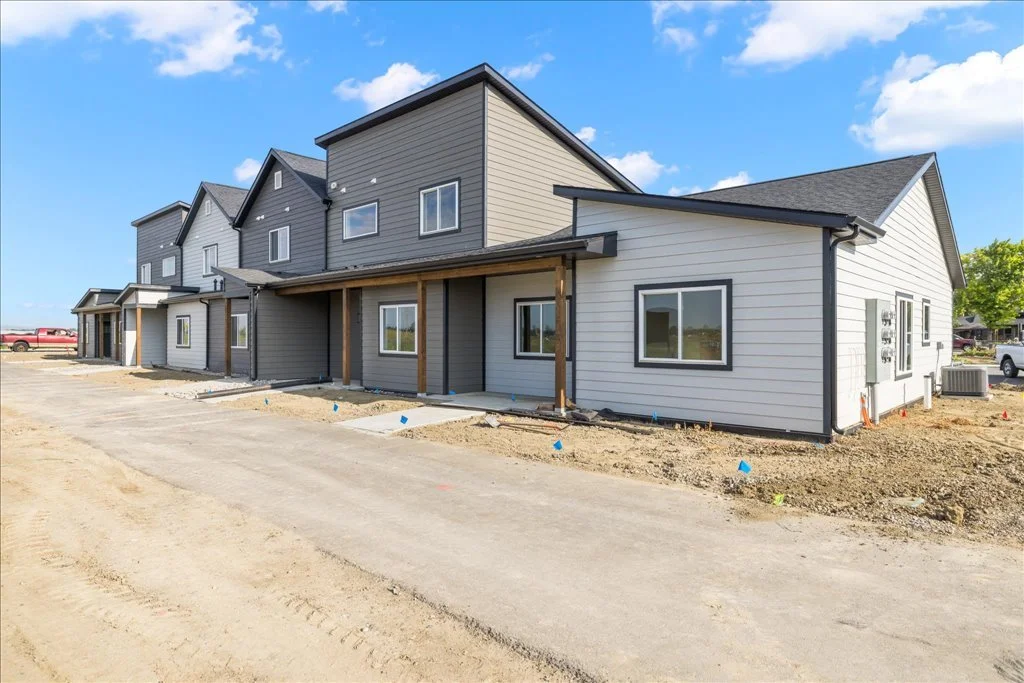 Newly constructed row of townhouses with gray and white siding, large windows, and small covered patios, under a partly cloudy sky.