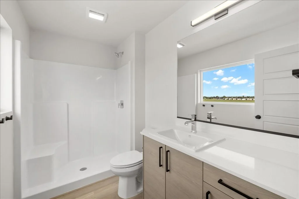 Modern bathroom with a shower, toilet, and vanity with a sink, large window with a view of a clear blue sky, and white walls.