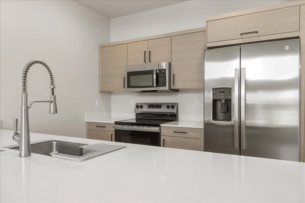 Modern kitchen with a white countertop, stainless steel appliances, light wood cabinets, and a pull-down kitchen faucet.