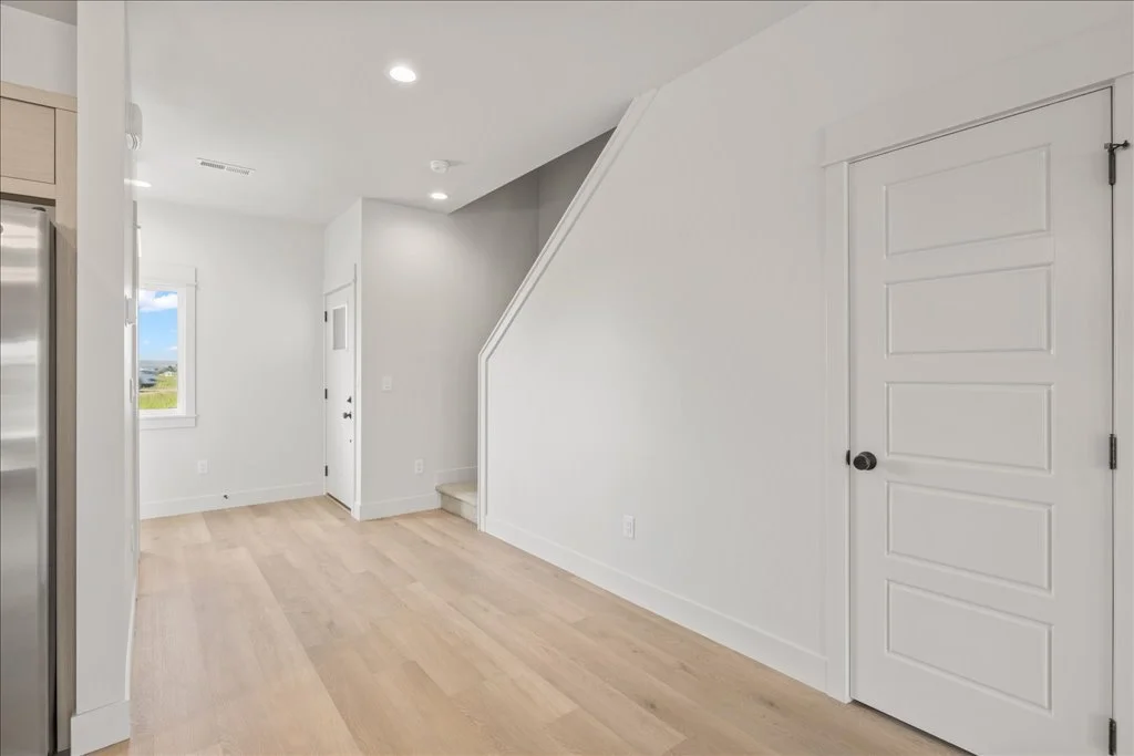 Interior of a modern home with white walls, light wood flooring, a staircase with a white banister, a closed white door, and a window showing an outdoor landscape.
