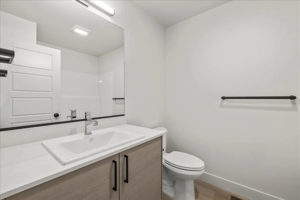 Bathroom with a white sink, wooden cabinet, and a toilet, with a large mirror above the sink and a black towel bar on the white wall.