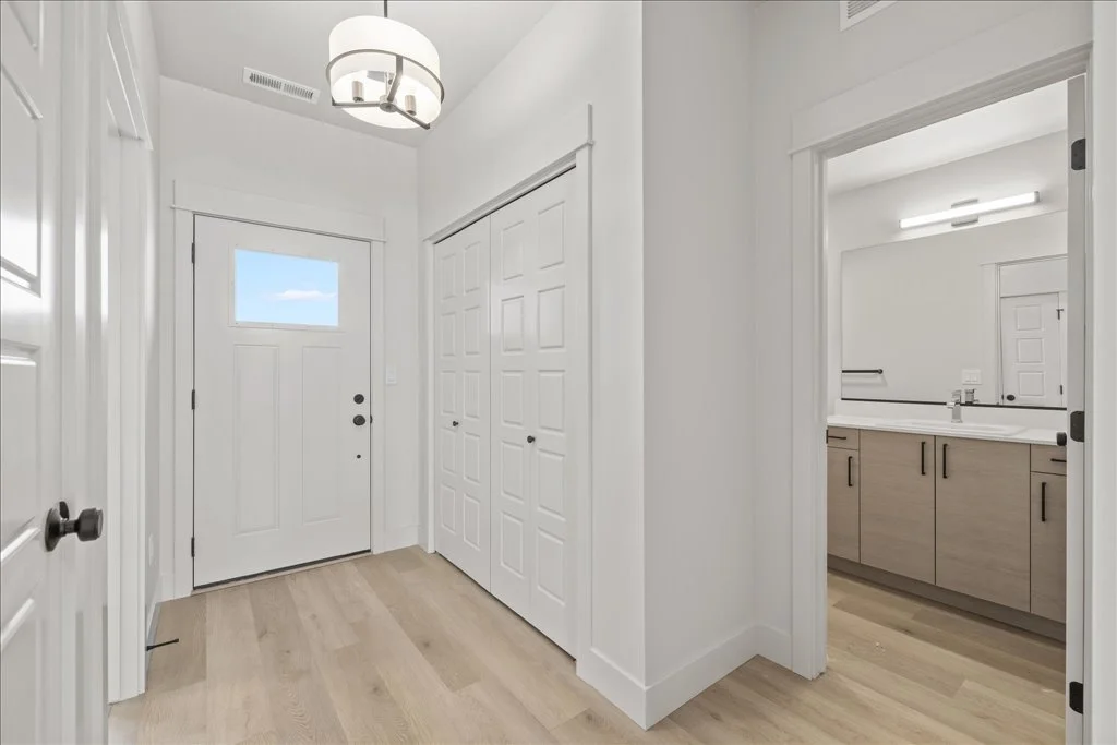 Interior view of a modern home's entryway and bathroom with white walls, light wooden flooring, a white front door with window, closet doors, and a bathroom with beige cabinetry, white countertop, and large mirror.