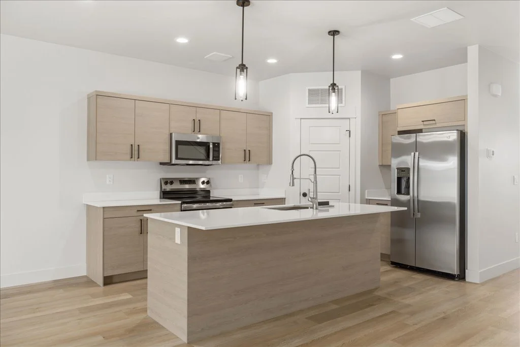 Modern kitchen with light wood cabinets, white countertops, stainless steel appliances, and pendant lighting.