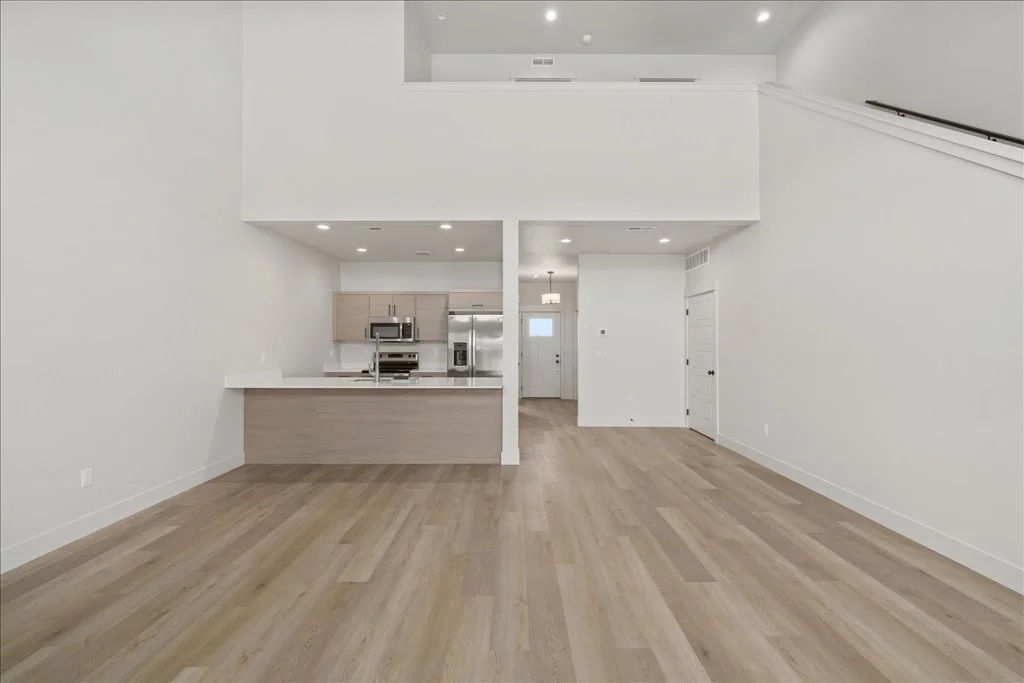 Empty modern open-concept living room and kitchen with white walls, light wood flooring, and stainless steel appliances.