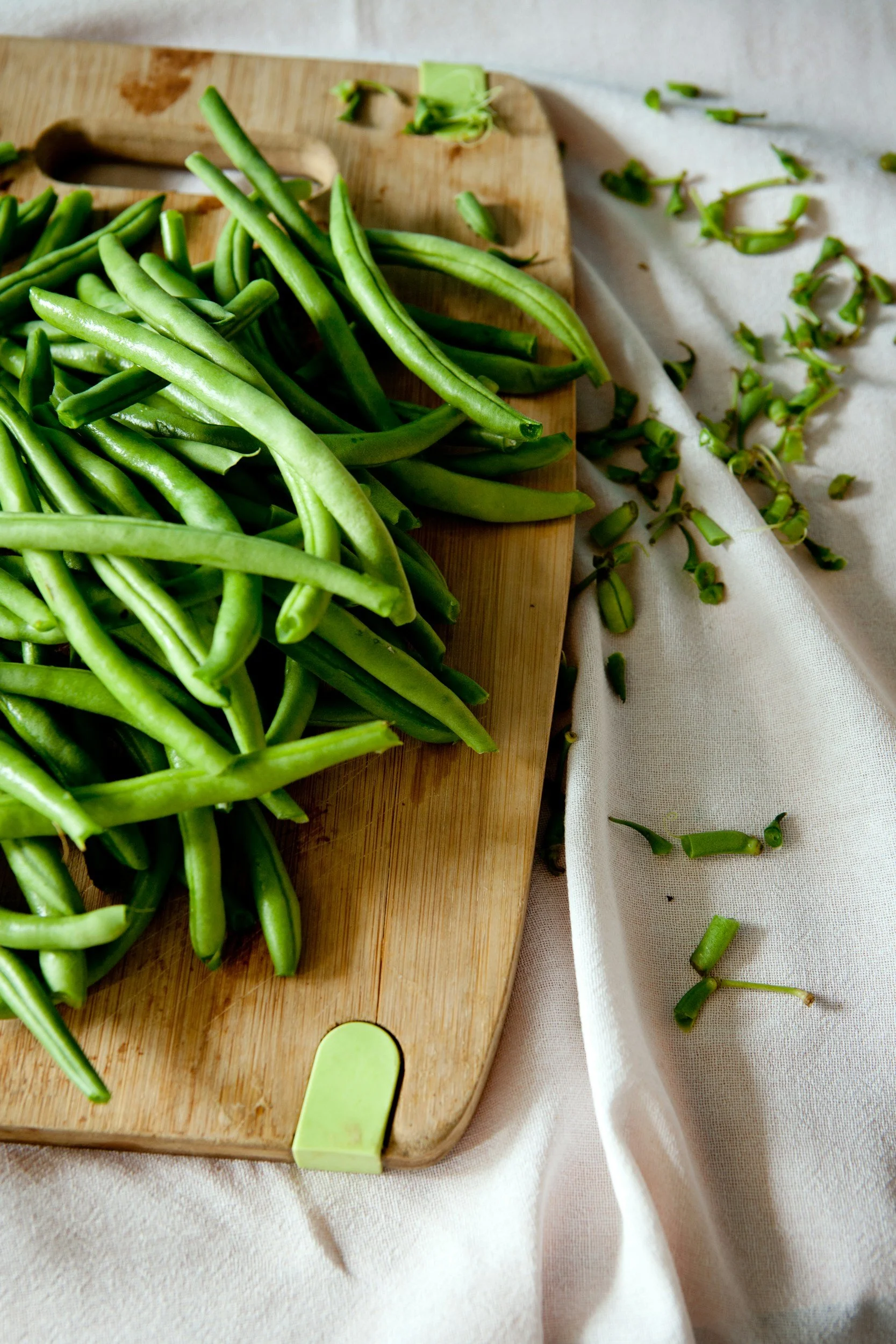 Fresh green beans on a wooden cutting board with chopped beans and bean trimmings on a white cloth.