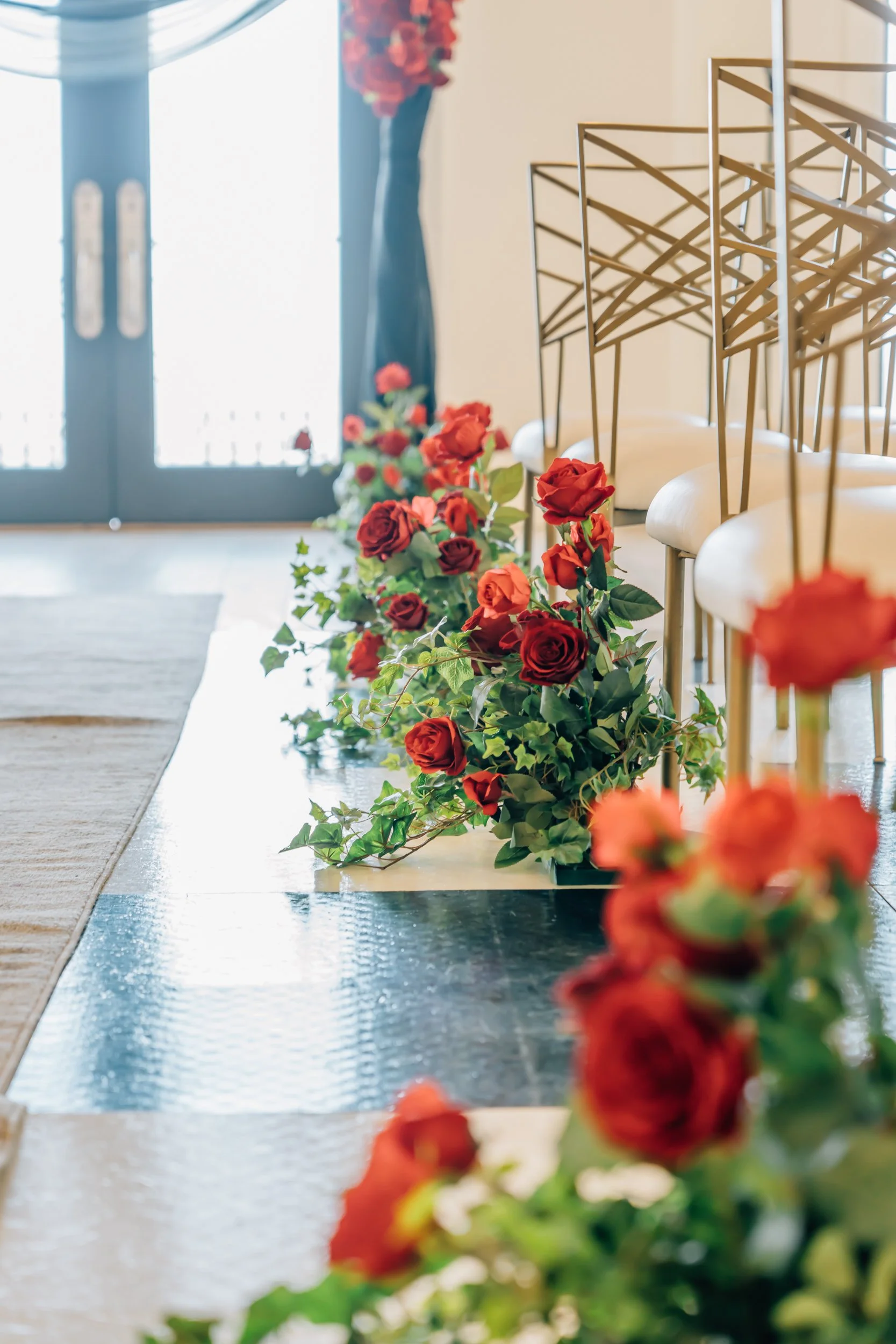A wedding or event ceremony setup with a row of red and orange roses and green foliage along the aisle, with chairs and a glass door in the background.