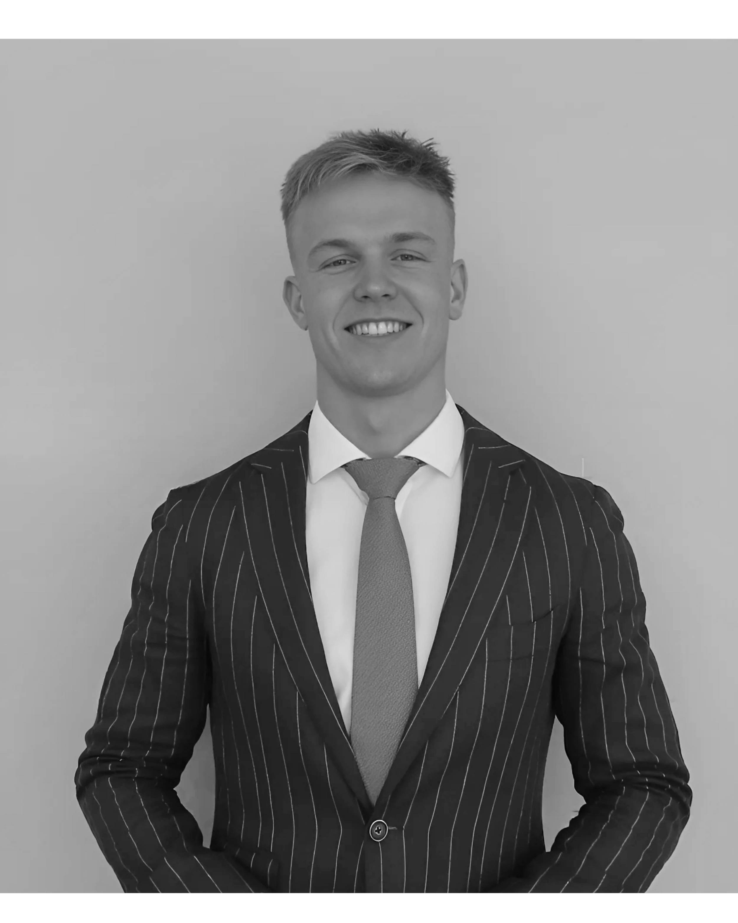 Black and white photo of a young man in a pinstripe suit with a tie, smiling, standing against a plain background.