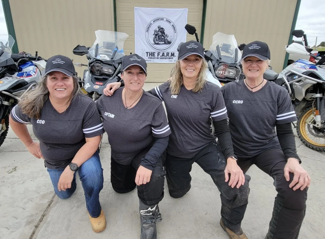 Four women posing in front of motorcycles, all wearing black shirts and caps with names and titles, smiling at the camera.