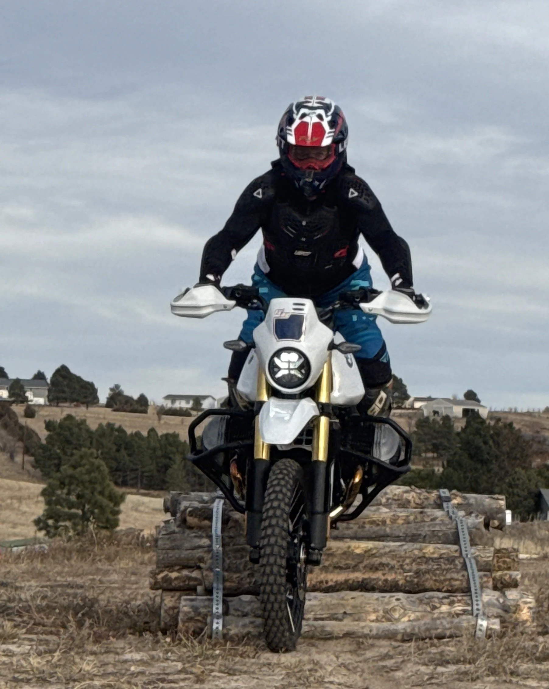 Motorcycle rider on a dirt trail with a log obstacle, wearing a helmet and riding gear, during daytime under a cloudy sky.