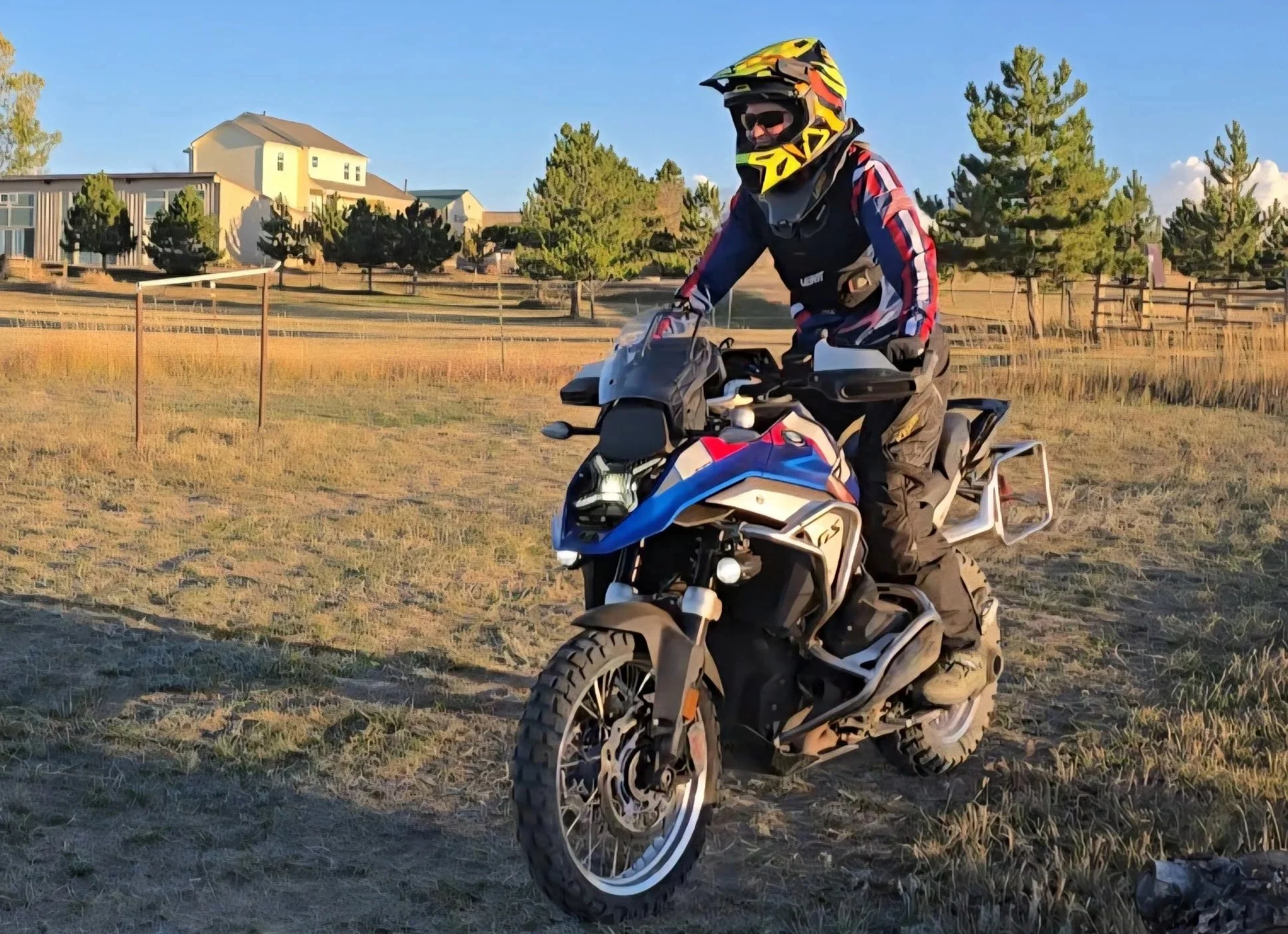 Person wearing a helmet, goggles, and riding gear on an adventure motorcycle, on grassy terrain with trees and houses in the background during sunset.