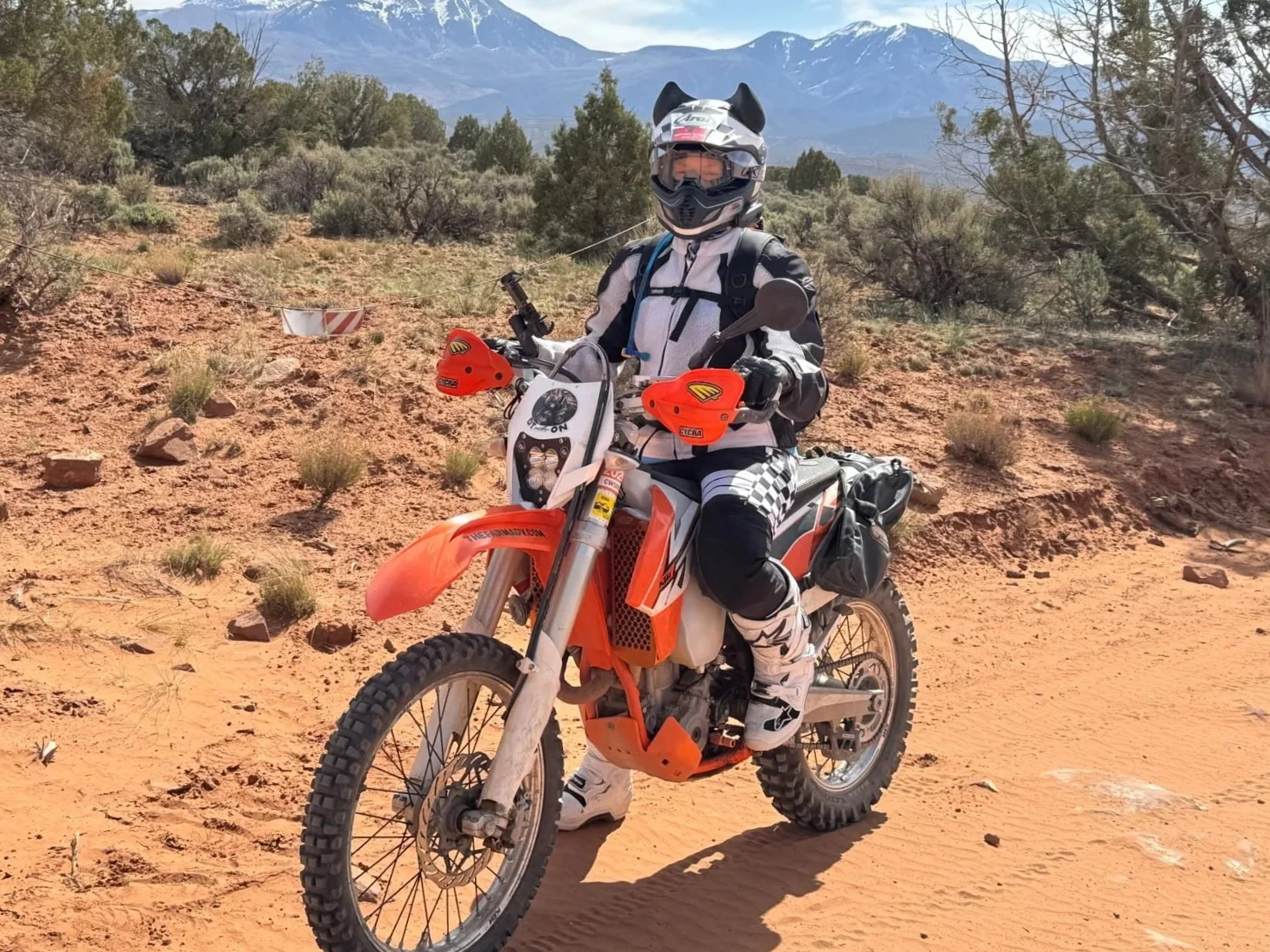 Motorcyclist wearing a helmet and riding gear on an orange off-road motorcycle in a desert landscape with mountains in the background.