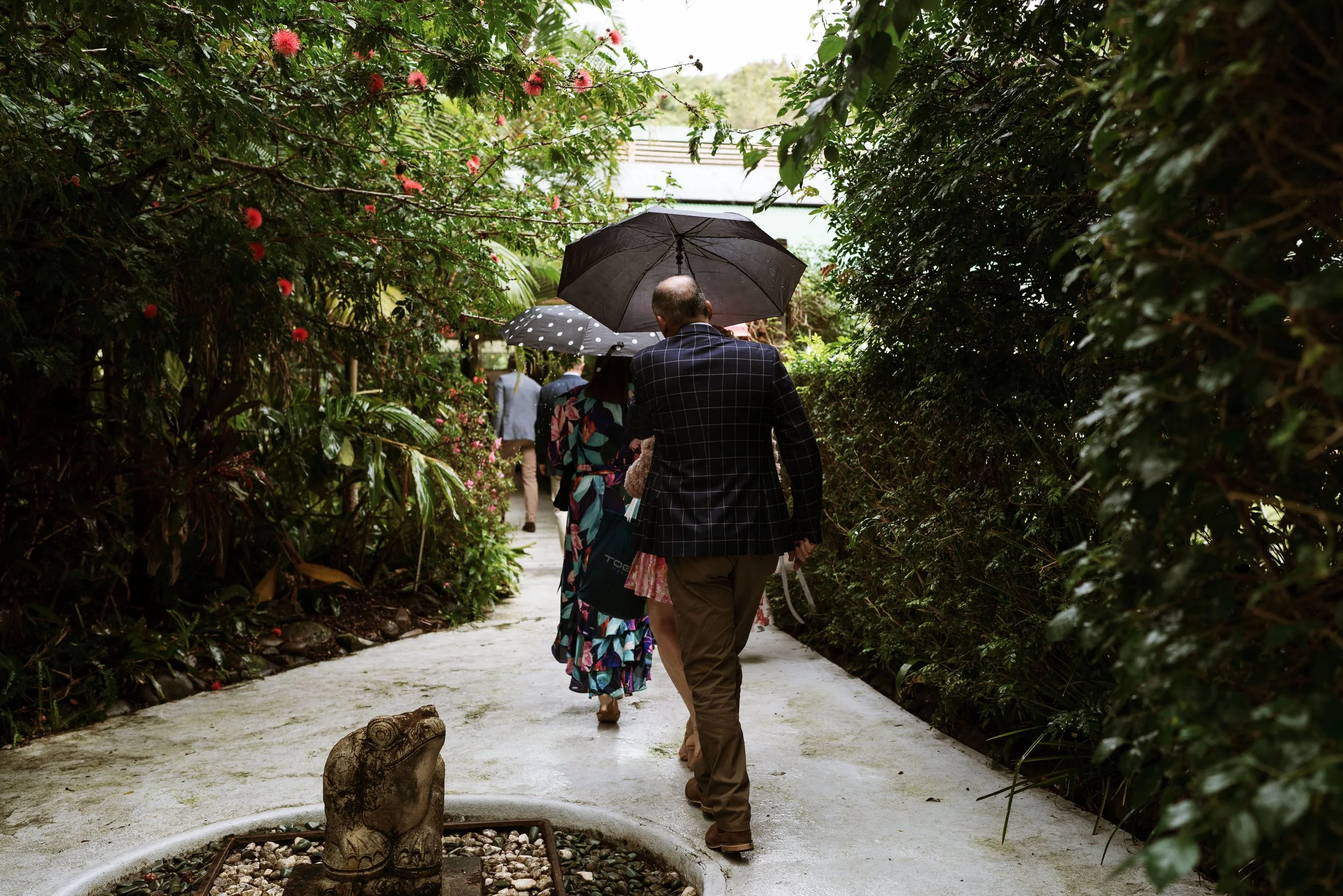 People walking along a garden path holding umbrellas, surrounded by lush green bushes and flowers.
