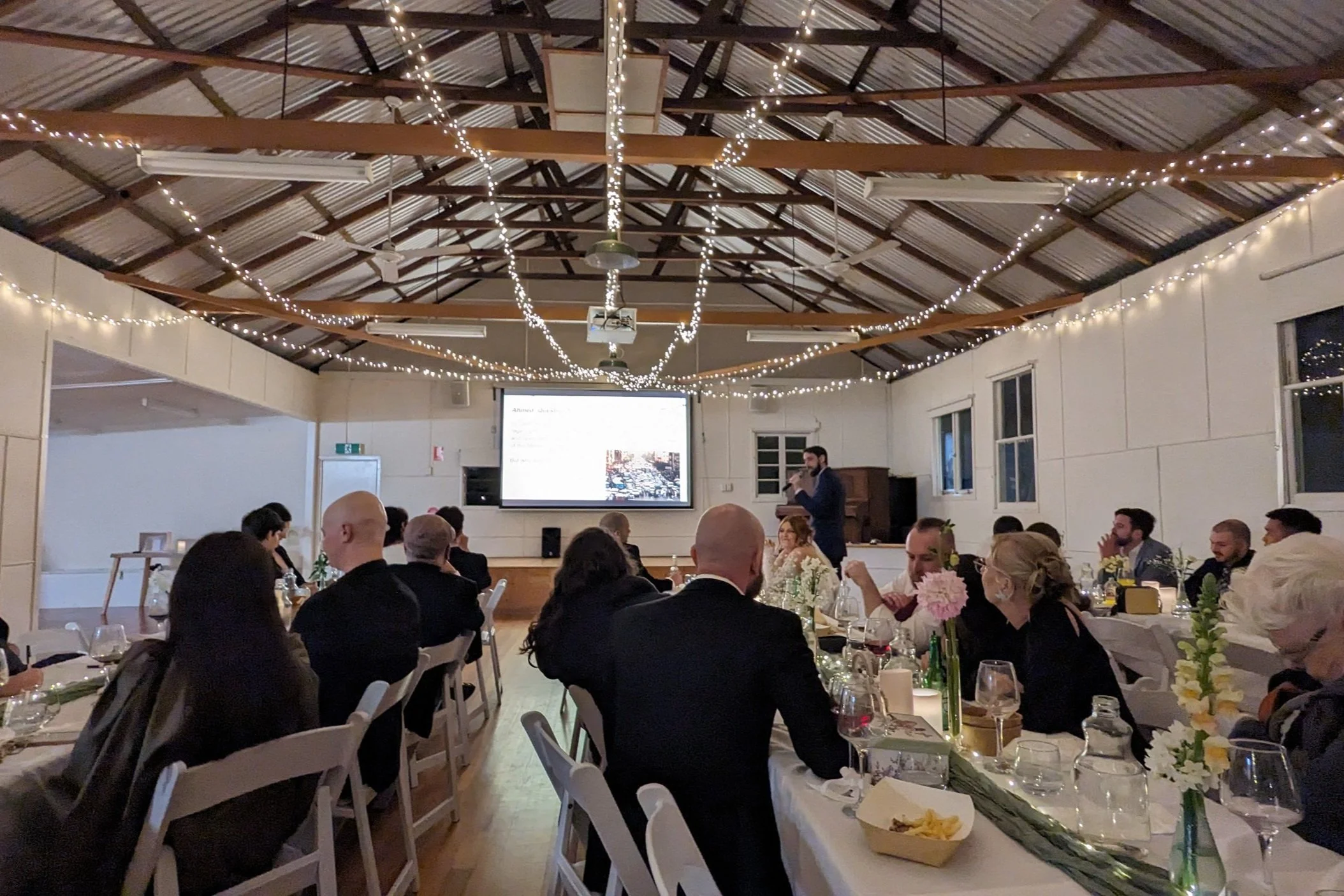 Indoor event with seated guests, a speaker at the front near a large screen, and string lights hanging from the ceiling.