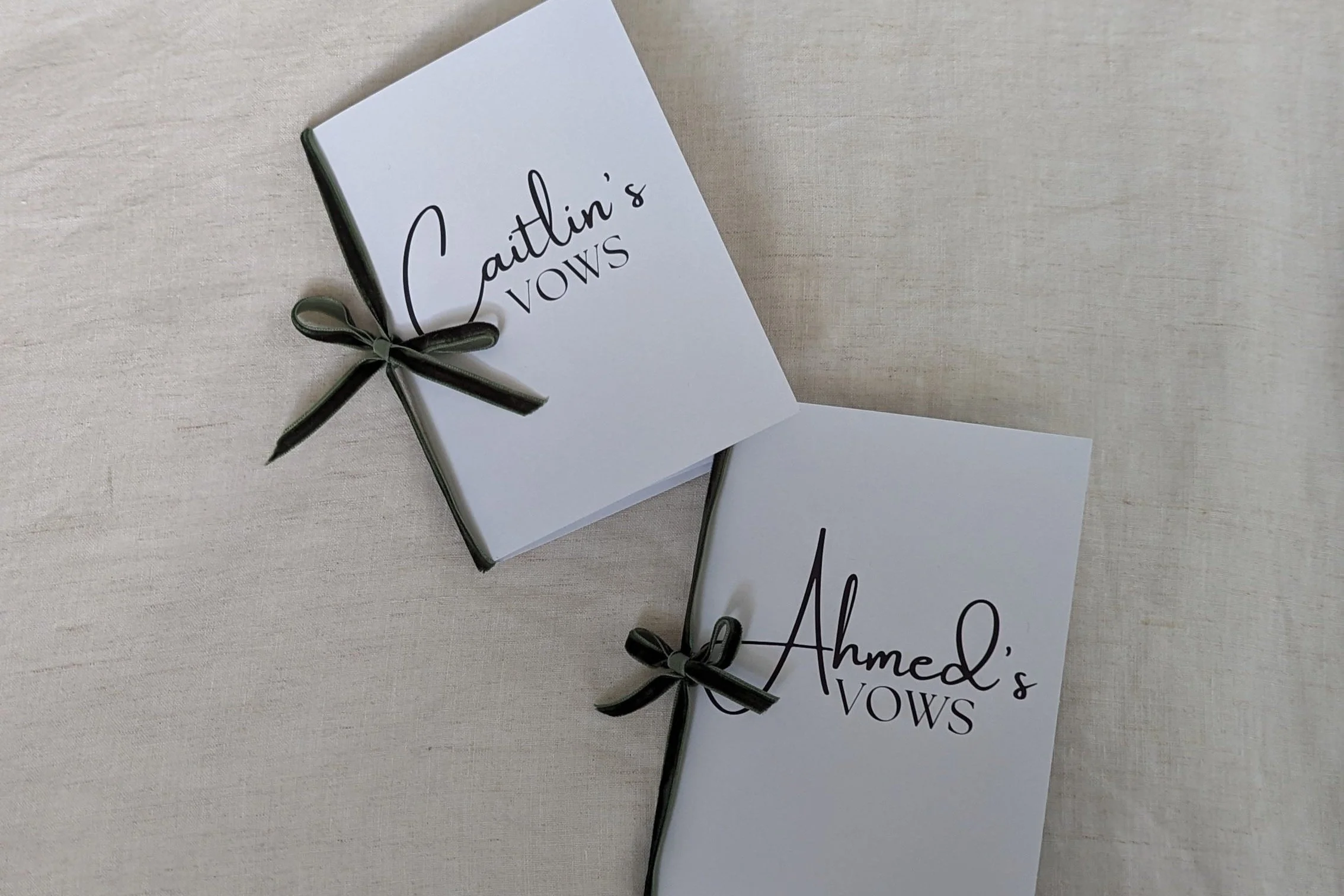 Two white vow books, one labeled 'Caitlin's VOWS' and the other labeled 'Ahmed's VOWS', each with black bows, placed on a light-colored fabric surface.