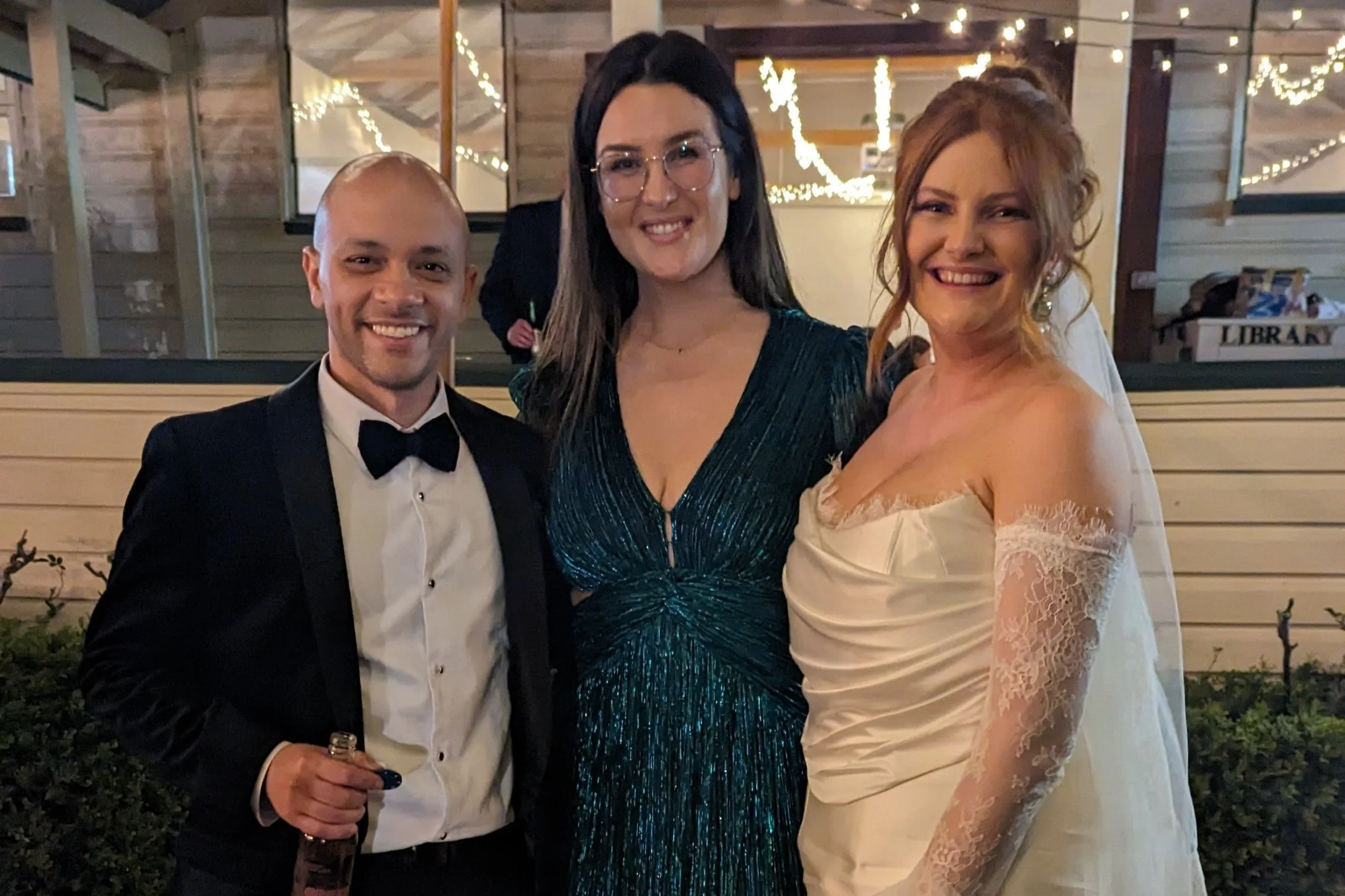 Three friends dressed in formal attire at a celebration. The man on the left is in a tuxedo holding a drink, the woman in the middle is wearing a shimmering dark green dress with glasses, and the woman on the right is in a wedding gown with lace slee