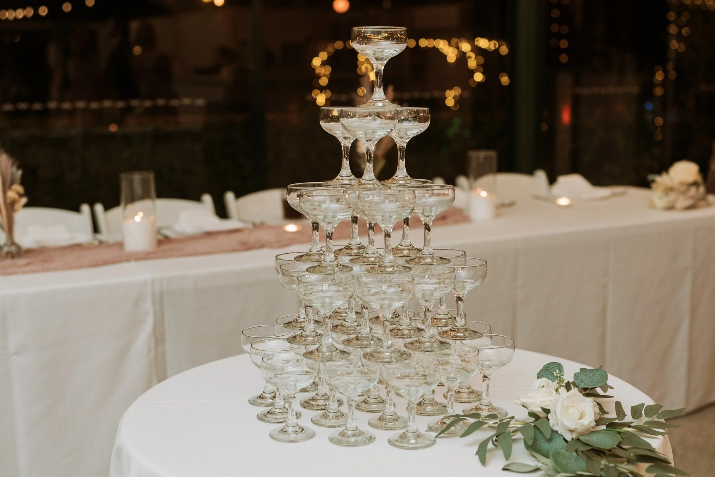 A pyramid of empty champagne glasses arranged on a round table, decorated with white flowers and greenery, at a celebration or event.