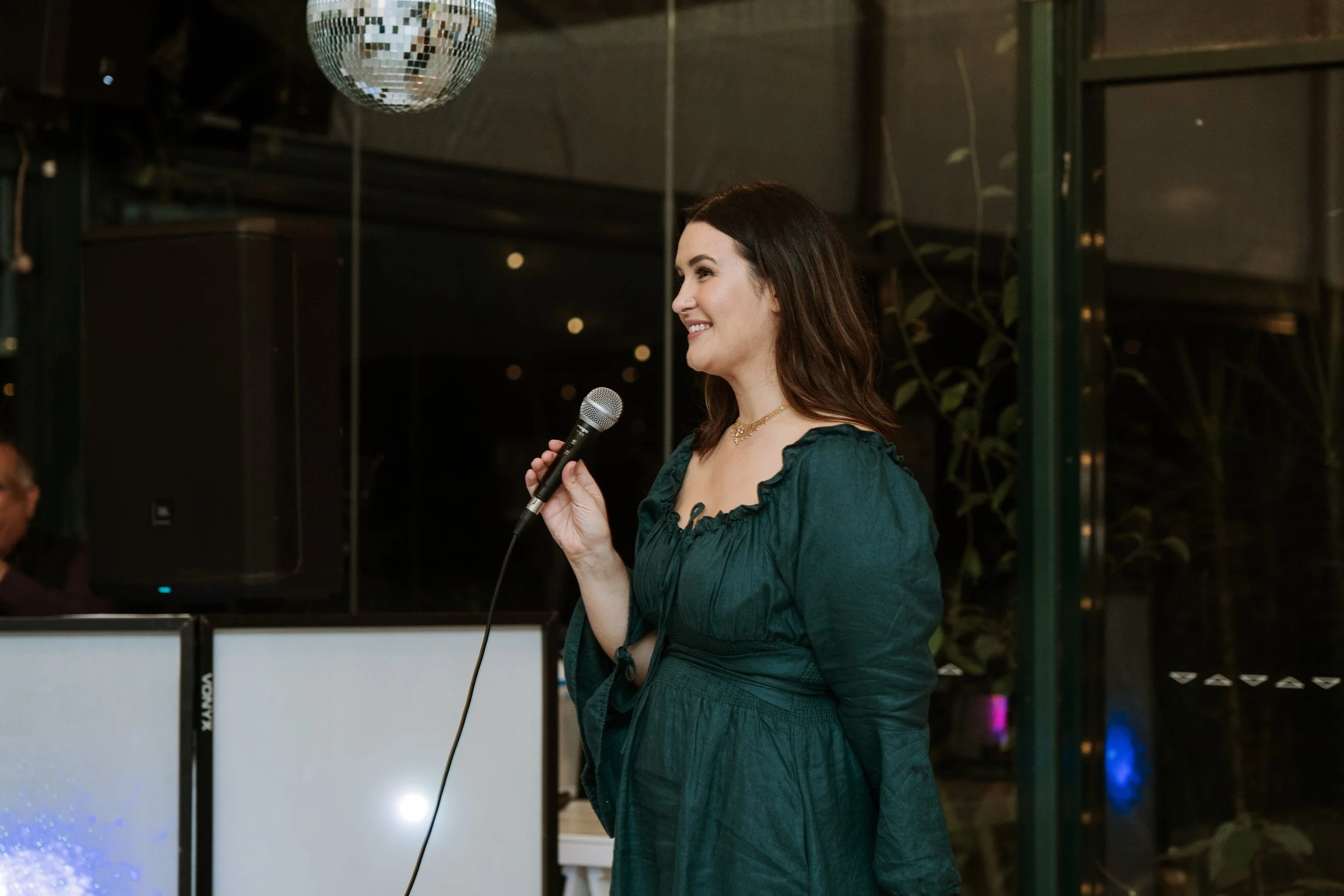 A woman in a dark green dress holding a microphone, smiling, with a disco ball above her.