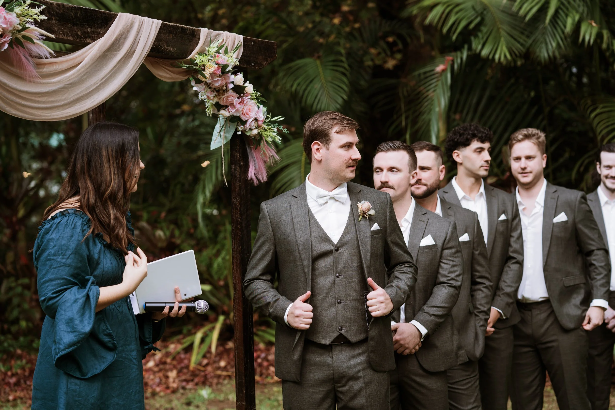 Groom and groomsmen standing outdoors at a wedding ceremony, with a woman officiant holding a microphone and a floral arch in the background.
