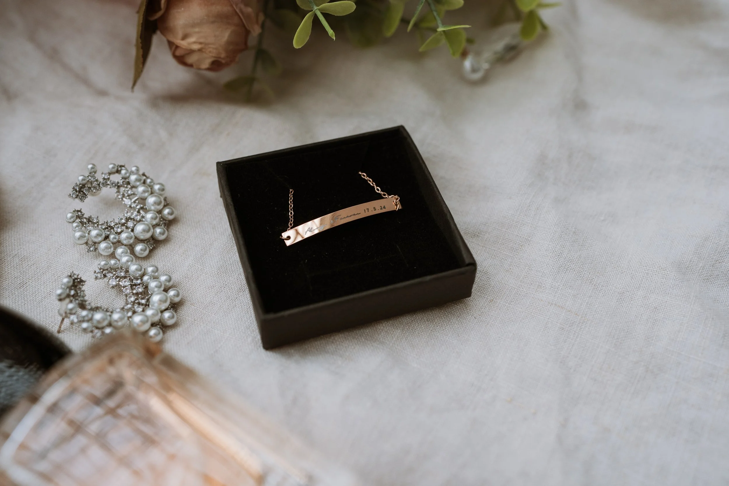 A black jewelry box holding a rose gold necklace with engraved message, beside a pair of pearl and rhinestone earrings on a white fabric surface.