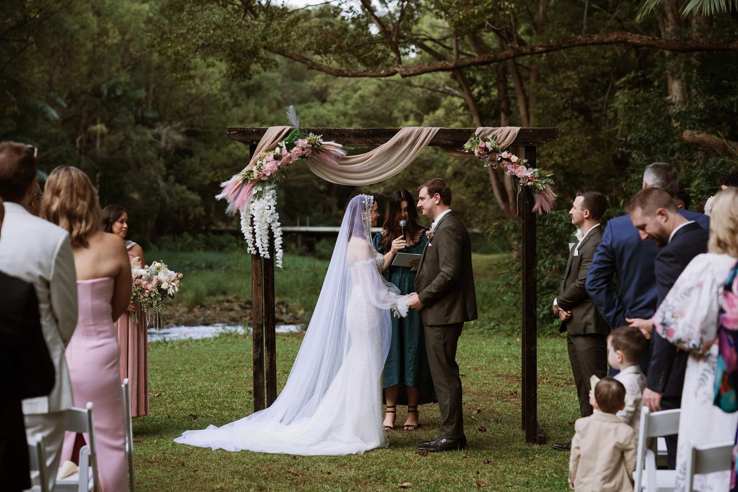 A wedding ceremony outdoors at a wooden arch decorated with pink and white flowers and fabric, with a bride and groom holding hands and facing each other. The bride is wearing a white wedding dress and veil, and the groom is in a gray suit. Guests ar