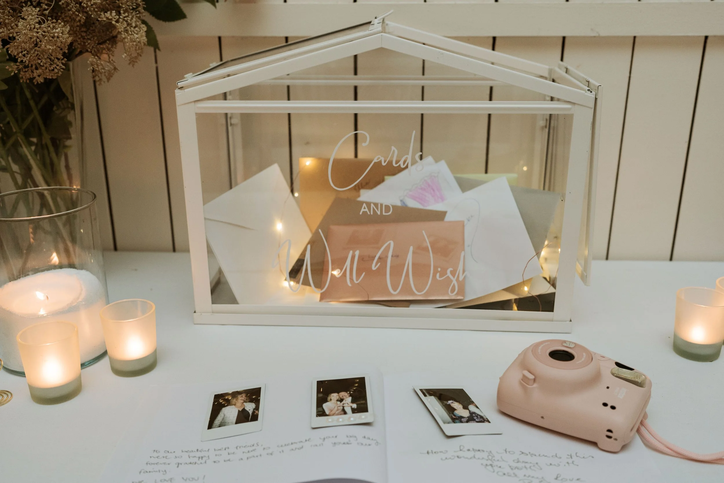 A glass terrarium with white frame labeled 'Cards and Wishing' filled with greeting cards and fairy lights, surrounded by candles, Polaroid photos, and a pink instant camera on a white table.