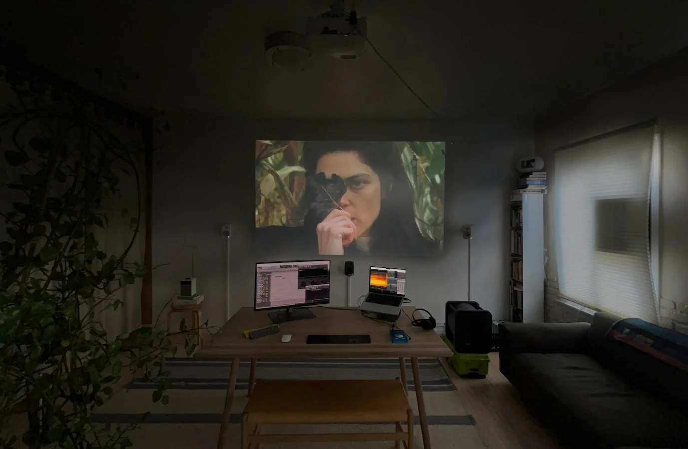 Living room with a large projection screen showing a woman holding a gun to her face, monitors on a desk, and a couch on the right, during a movie or show night.