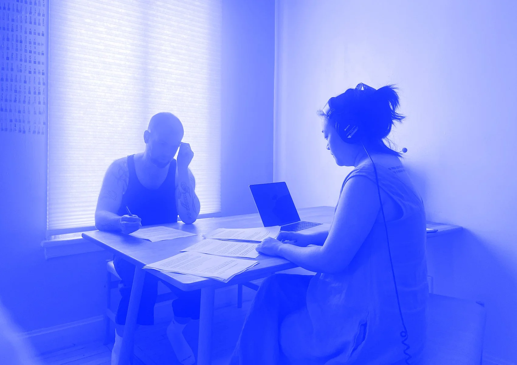 A woman wearing headphones sitting at a table with paperwork and a laptop, talking to a man across from her in a well-lit room with a window.