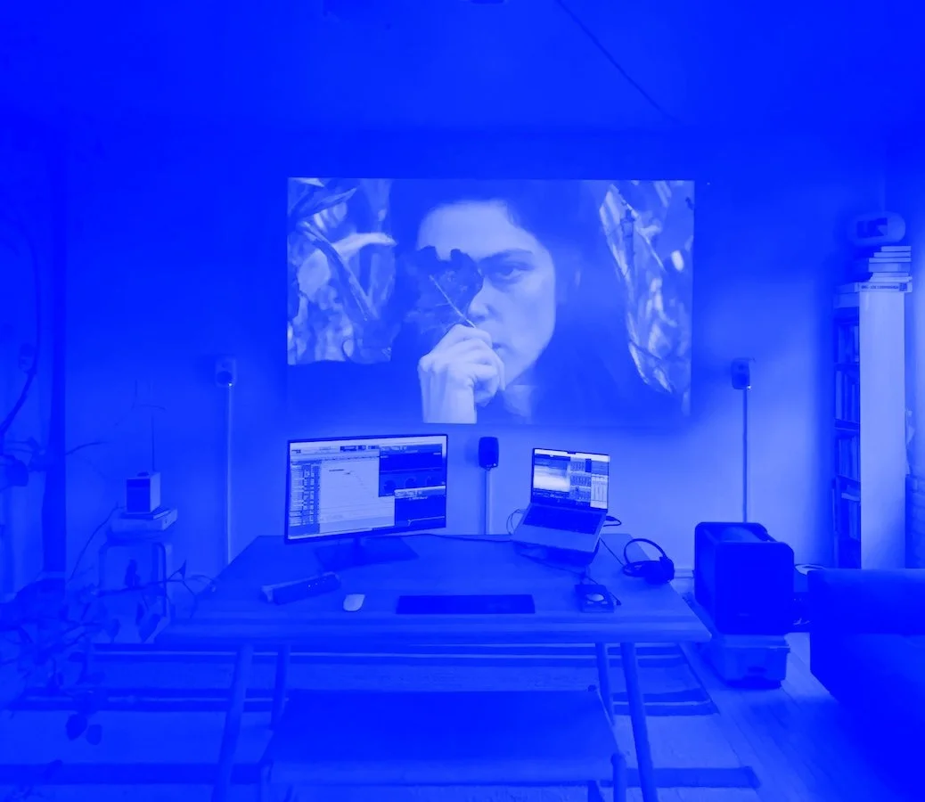 Home entertainment setup with a large wall projection of a woman holding a leaf, two computer monitors on a desk displaying digital audio workstations, speakers, and various electronic equipment in a room with blue lighting.