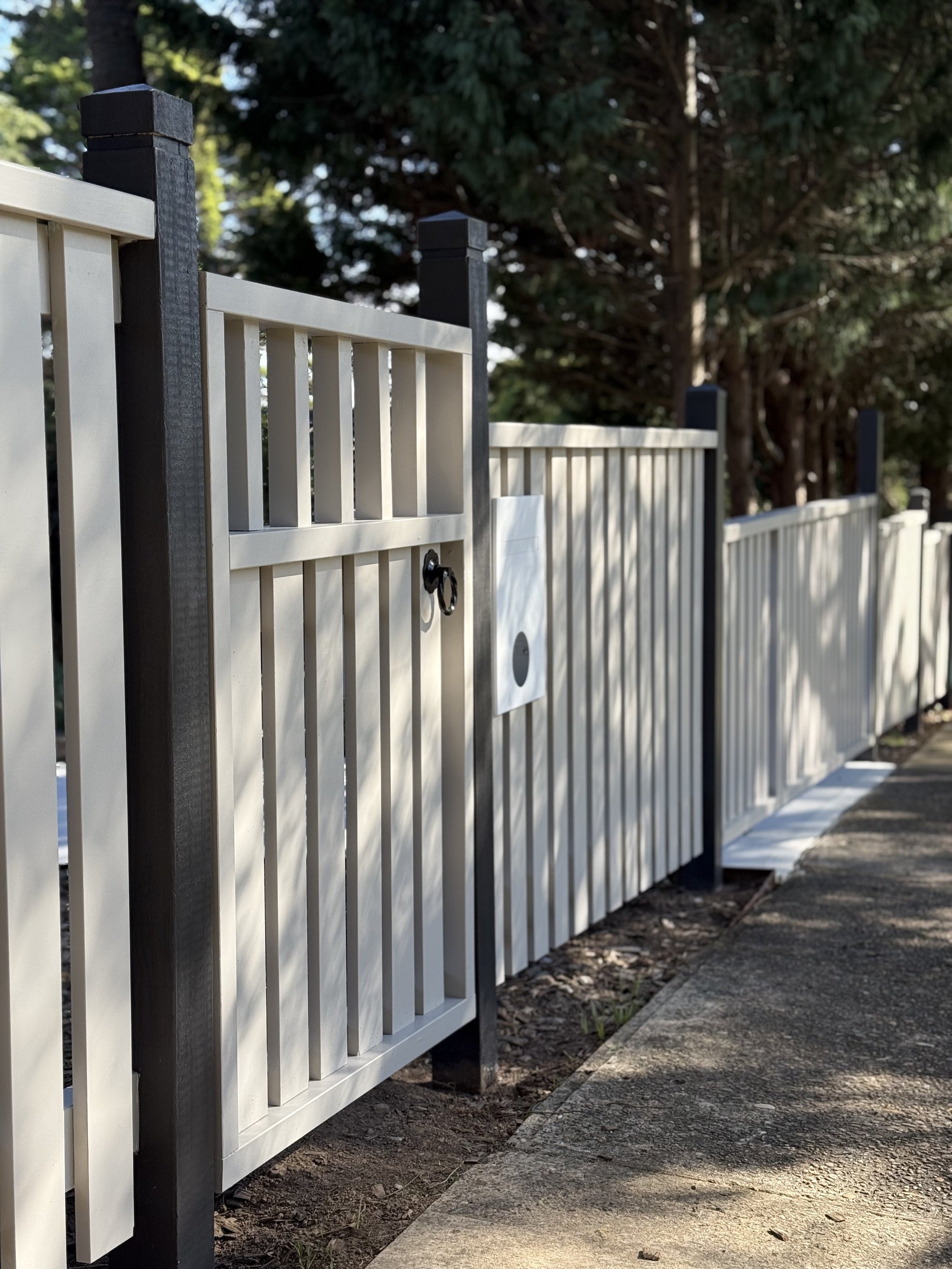 A white wooden fence with black posts and a small gate, installed alongside a sidewalk with trees in the background.