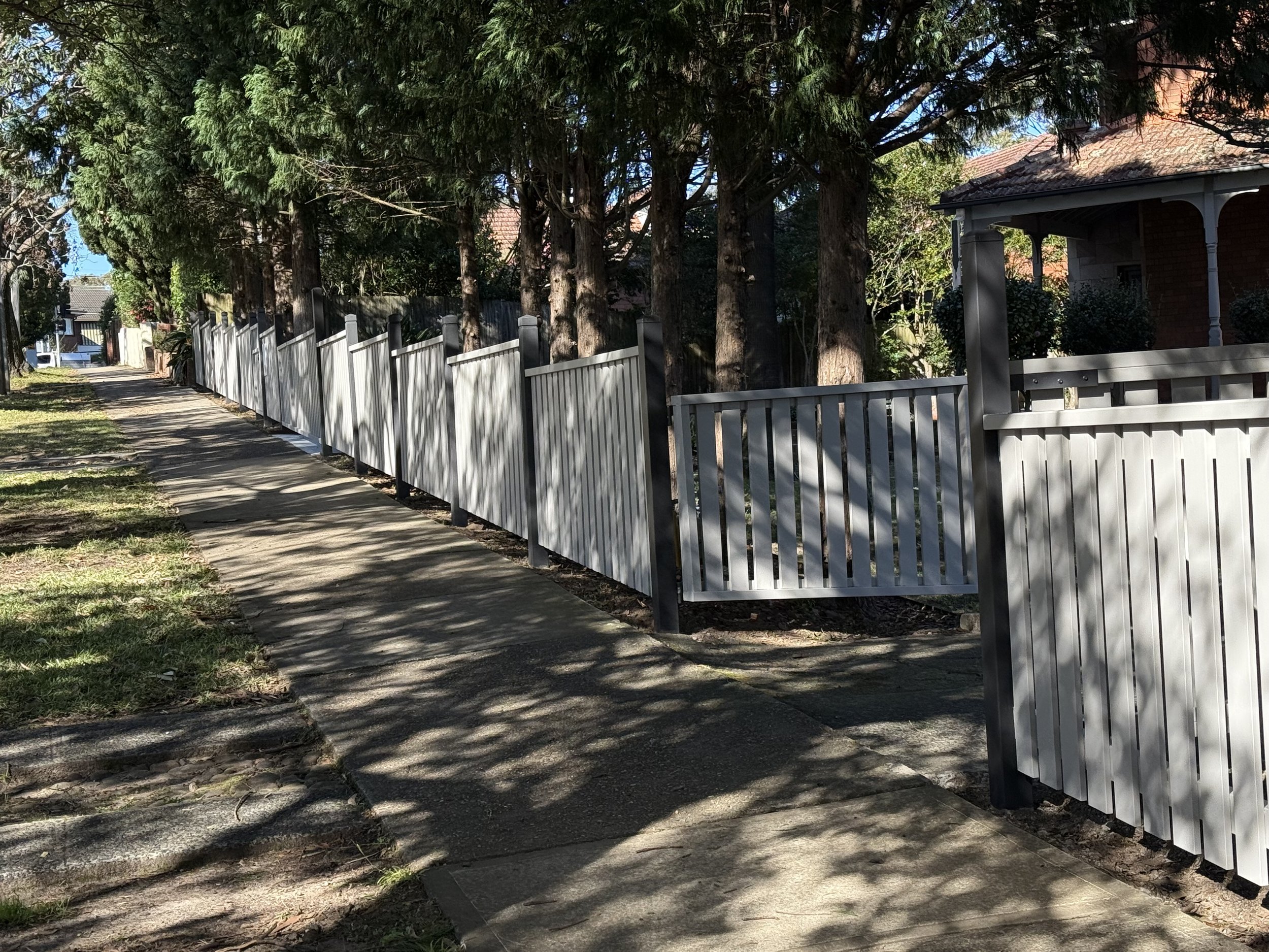 Residential sidewalk with a white picket fence and trees casting shadows on the sidewalk.