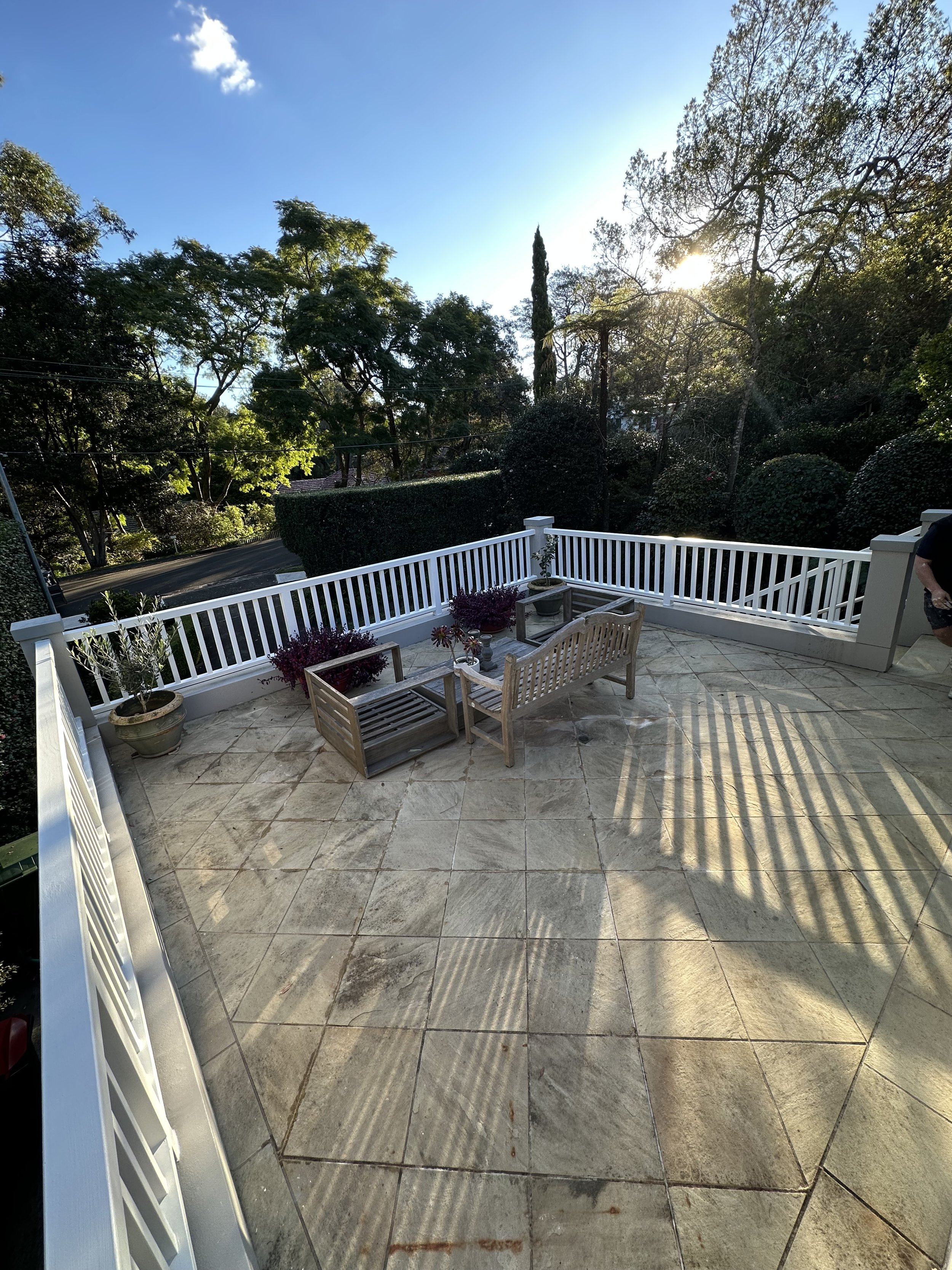 An outdoor patio with a tiled floor, white railing, wooden bench, two potted plants, and shaded by trees on a sunny day.