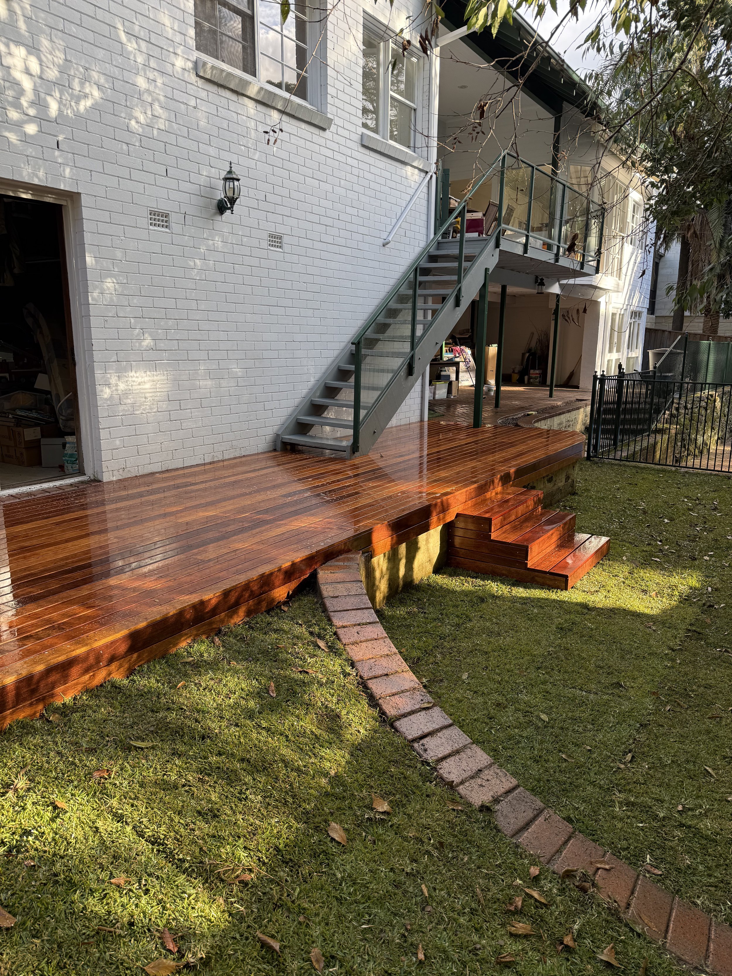 Backyard with a newly built polished wooden deck, brick stairs, and a white brick house with an exterior staircase. There is a small lawn with a curved brick pathway and some trees in the yard.