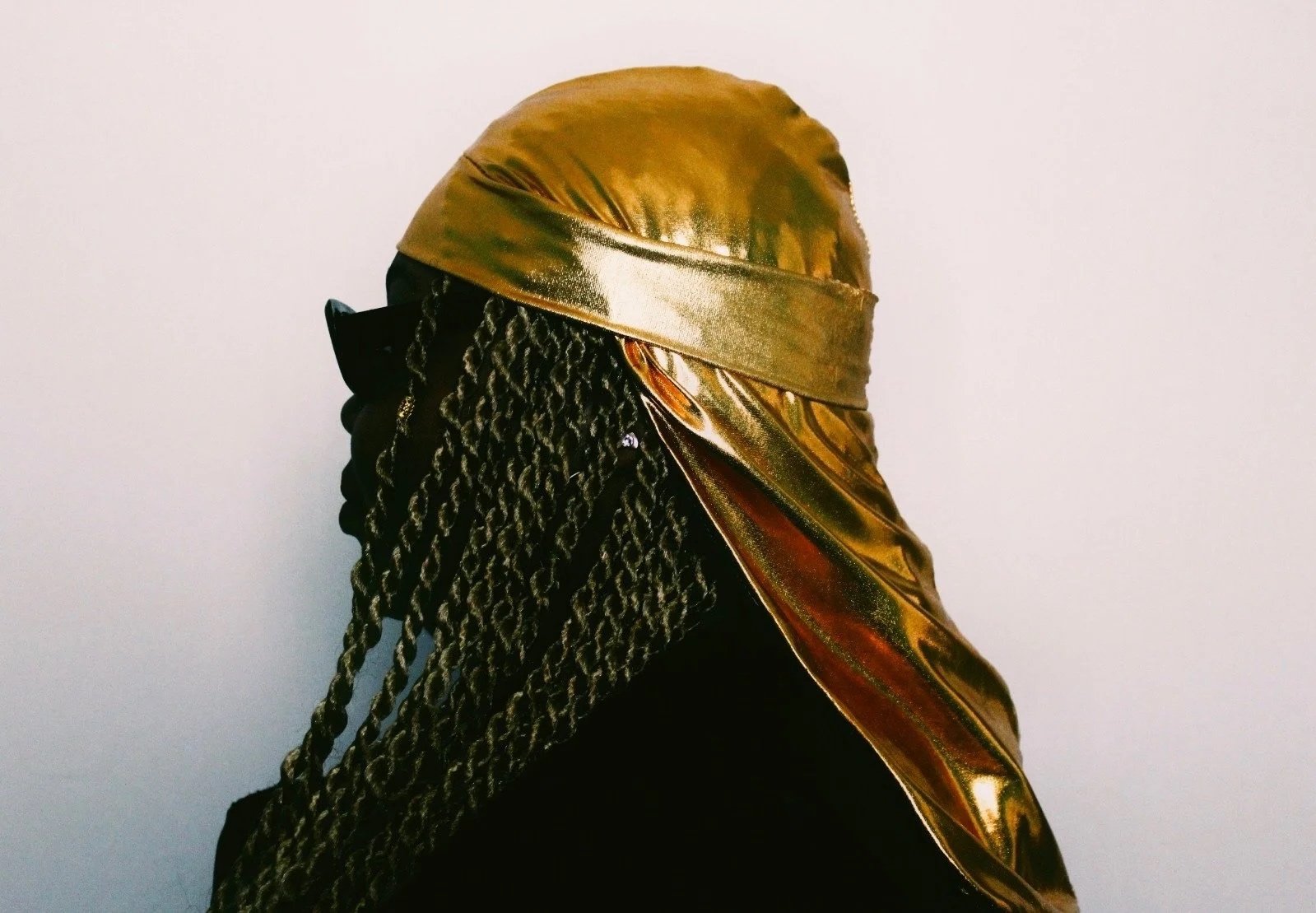 Person wearing a gold headwrap and gold silk-like scarf, with long braided hair, against a plain white background.