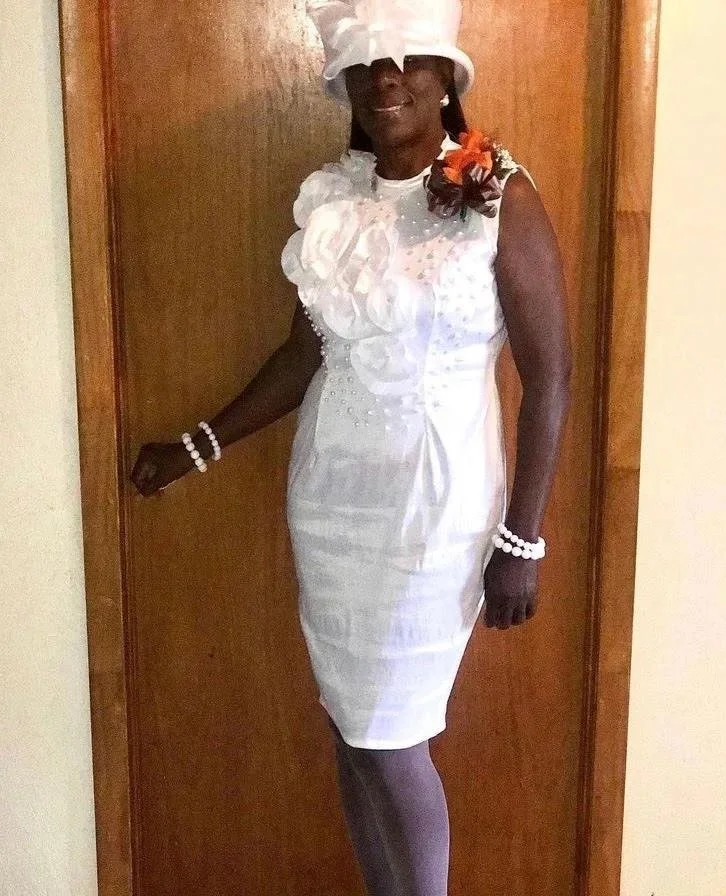 A woman in a white dress with a large floral decoration on the shoulder, wearing a hat, jewelry, and white shoes, standing indoors against a wooden door.
