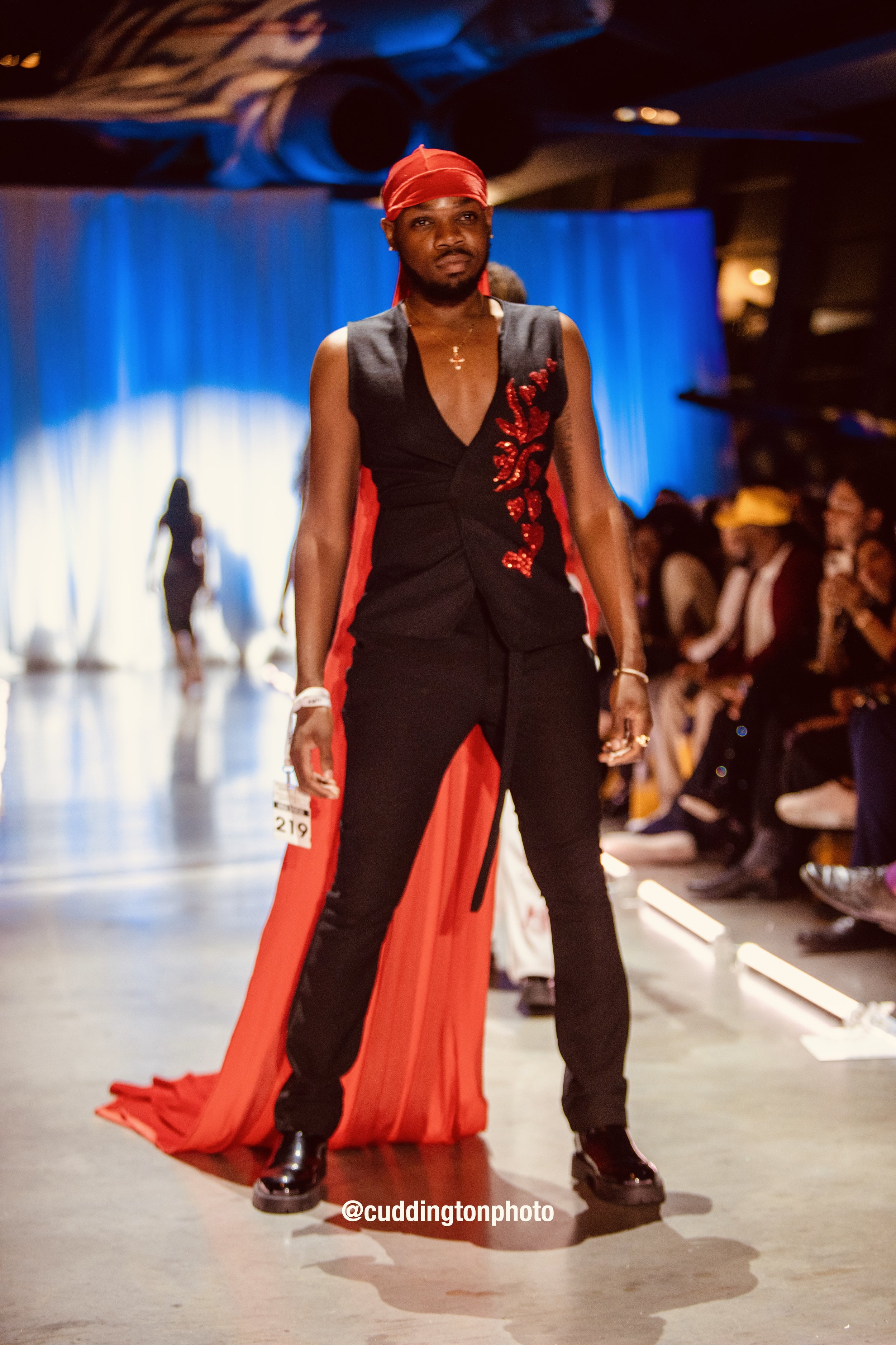 Person standing on runway at fashion show wearing black sleeveless outfit with red details and a long red cape, with audience seated on sides and a blue curtain background.