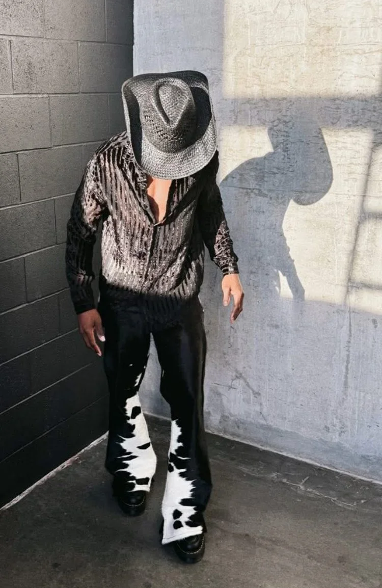 Person wearing a striped shirt, black pants with cow print on the legs, and a wide-brimmed hat, standing in a corner with a shadow cast on the wall.