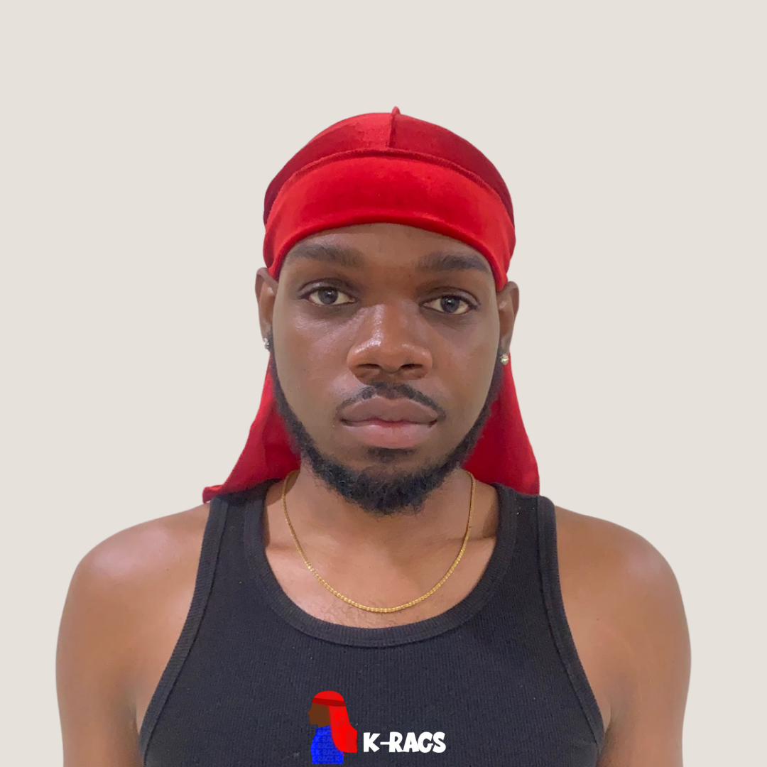 A young man with medium brown skin, wearing a red headscarf, black tank top with a graphic logo, gold necklace, and earrings, standing against a plain light background.