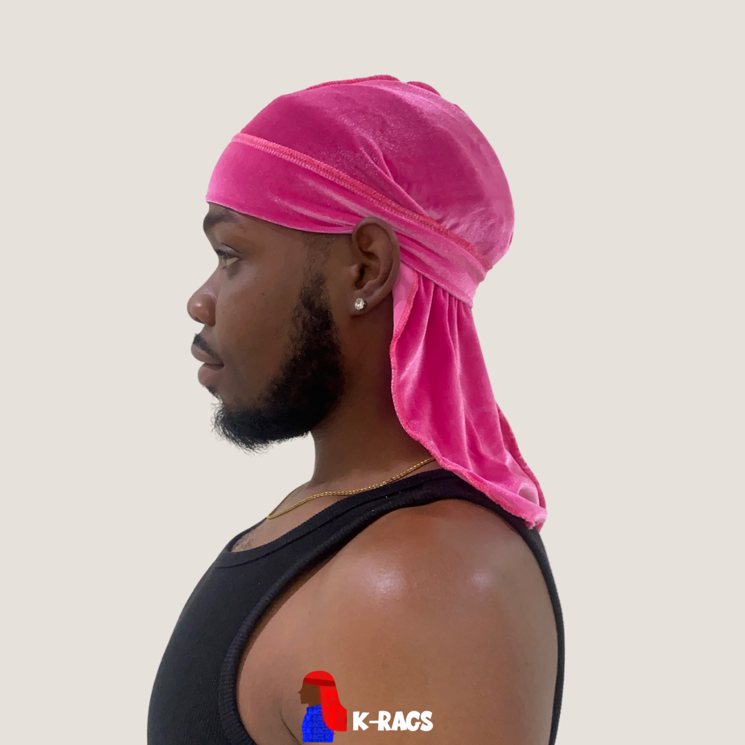 Profile of a young man wearing a bright pink satin durag, a black tank top, and a gold chain, standing against a plain, light background.
