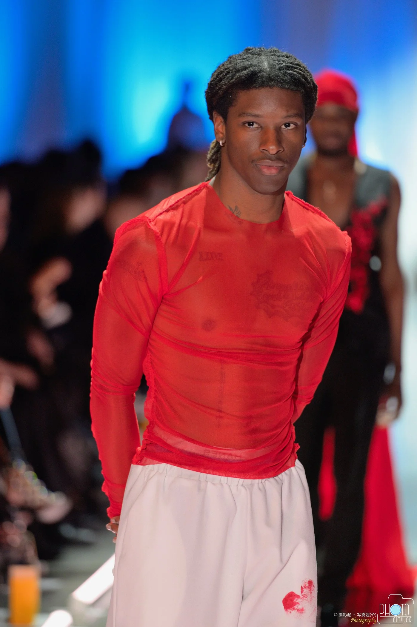 Male fashion model wearing a sheer red top and white pants walking runway at fashion show with audience in background.
