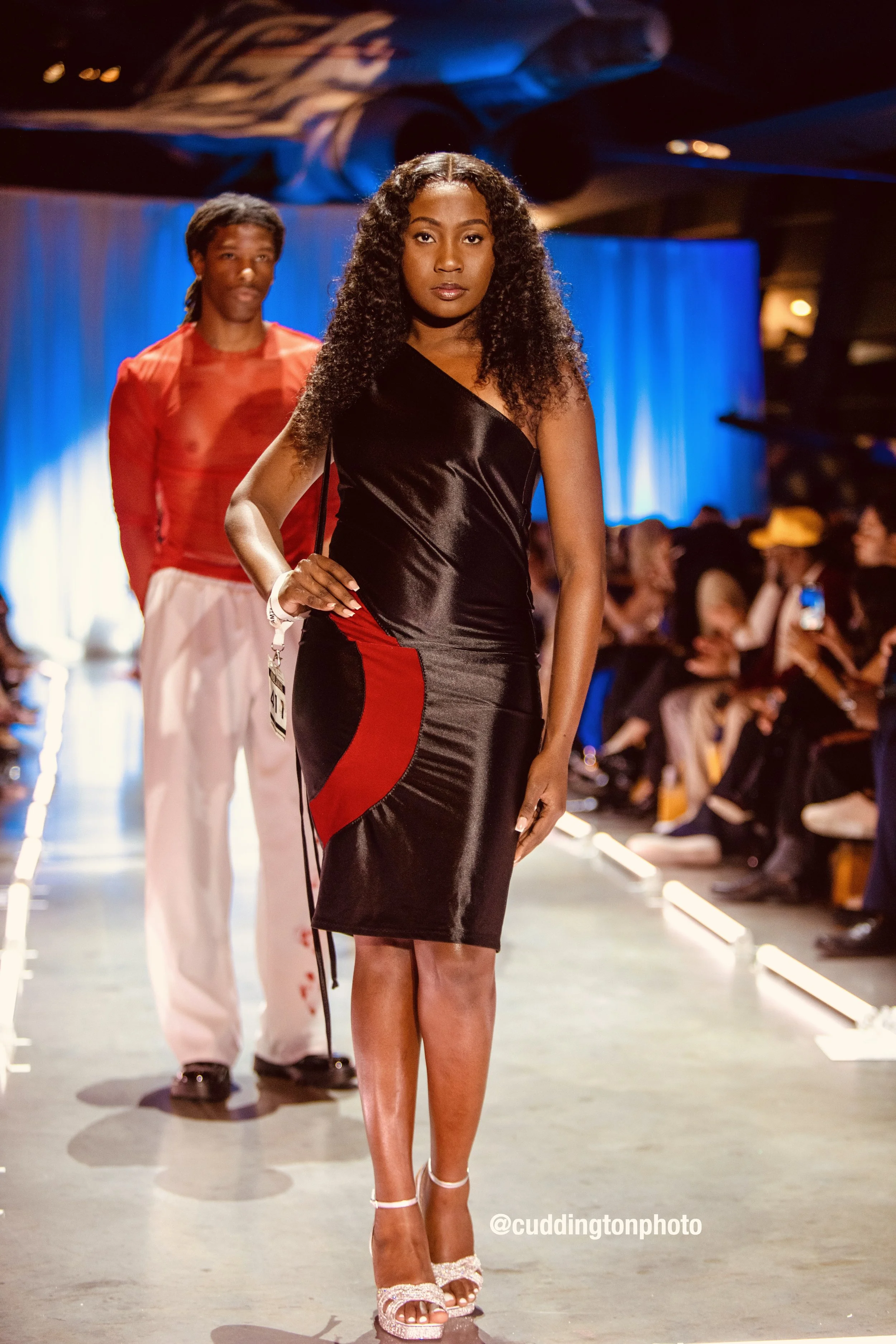 Model walking on runway in black and red dress, audience seated on sides, with person in red and white outfit behind.