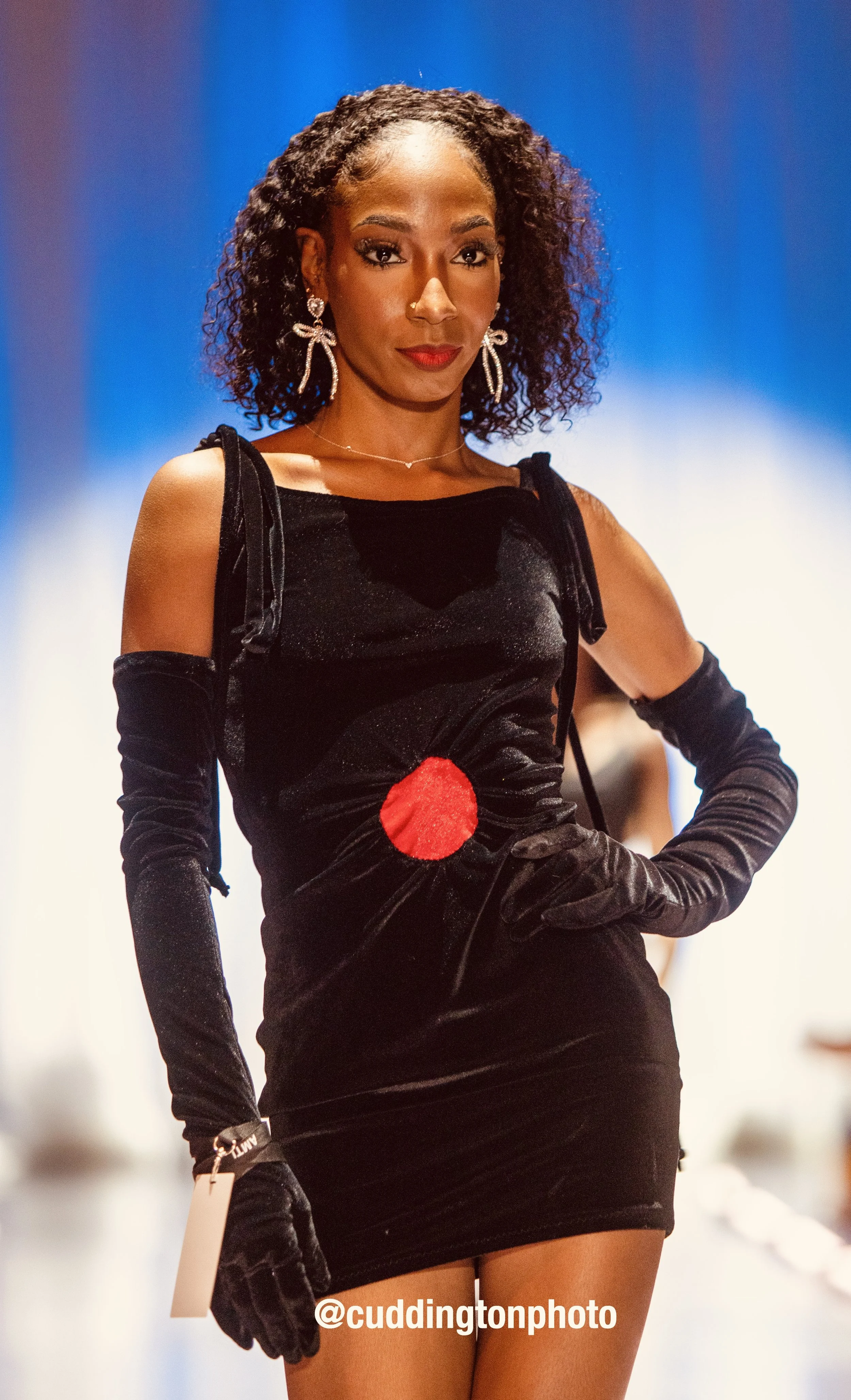A woman with curly hair on a runway, wearing a black dress with red circle detail, long black gloves, and statement earrings.