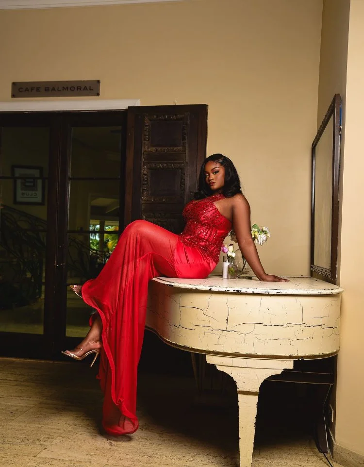 A woman in a red dress sitting on a vintage white piano in a room with a cream-colored wall, with a sign above the door reading 'Cafe Balmoral' and a mirror on the wall