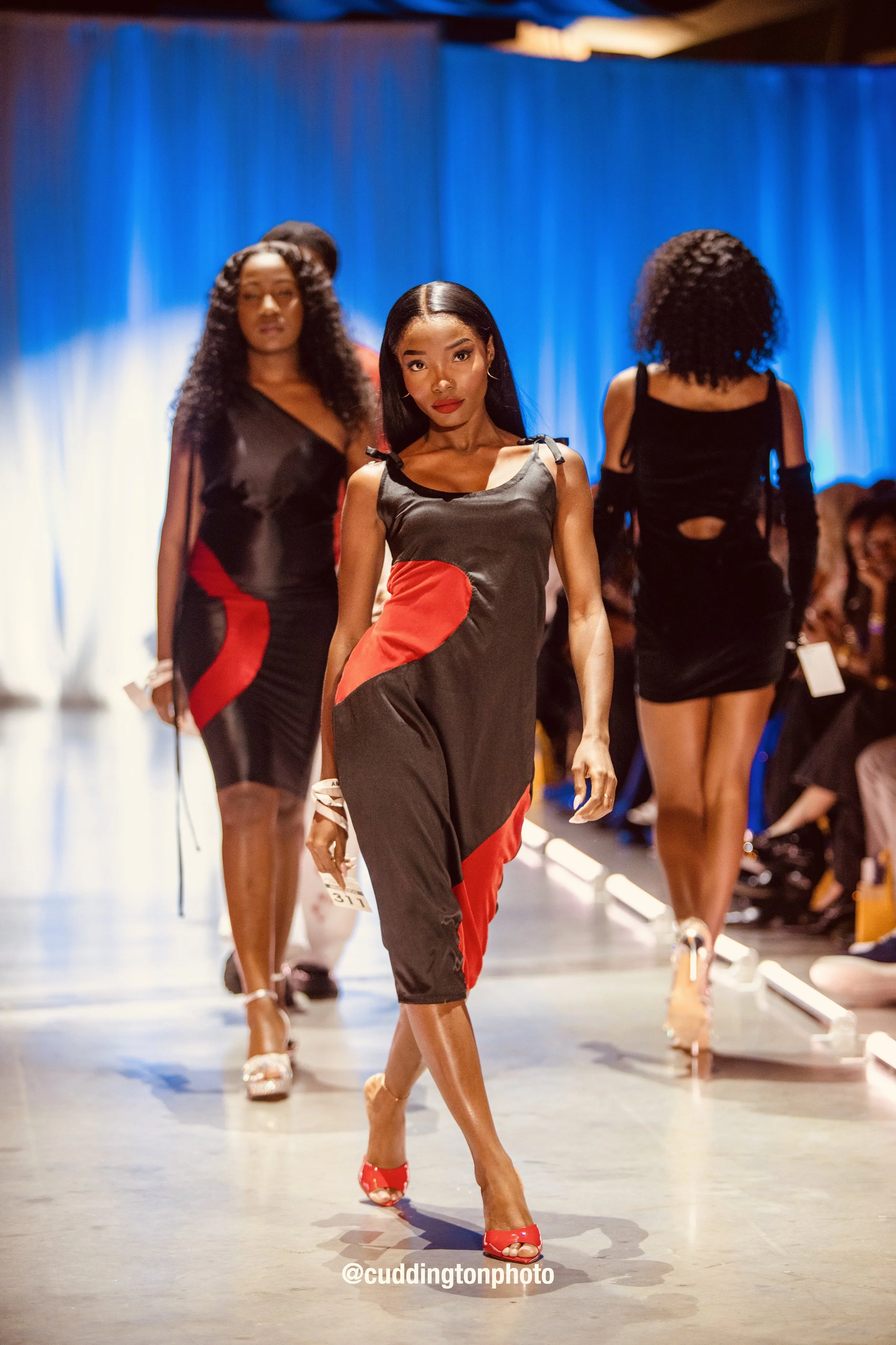 Models walking on a runway during a fashion show, wearing black and red dresses against a blue backdrop.
