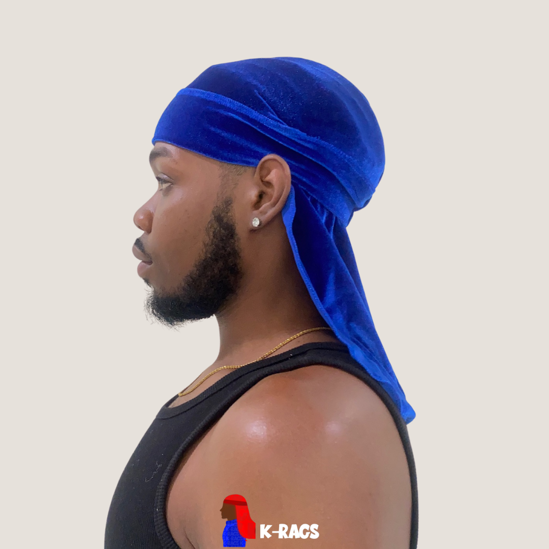 Profile view of a man with a beard, earrings, and a gold necklace, wearing a bright blue durag and a black tank top, with a neutral background.