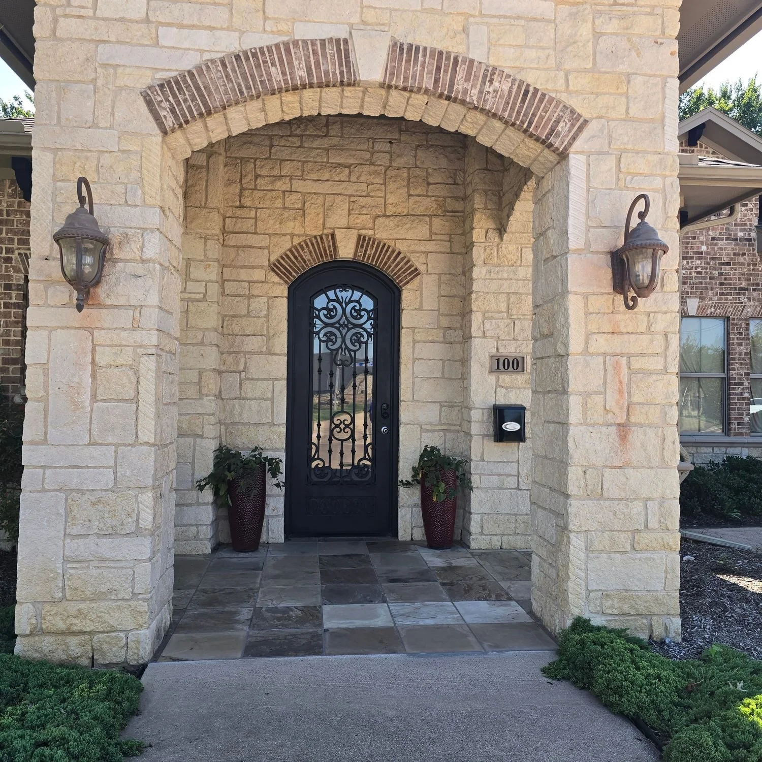 Front entrance of a house with a stone facade, black wrought iron door, two wall-mounted lanterns, potted plants, house number 100, and a mailbox, with a covered porch and garden plants in the foreground.