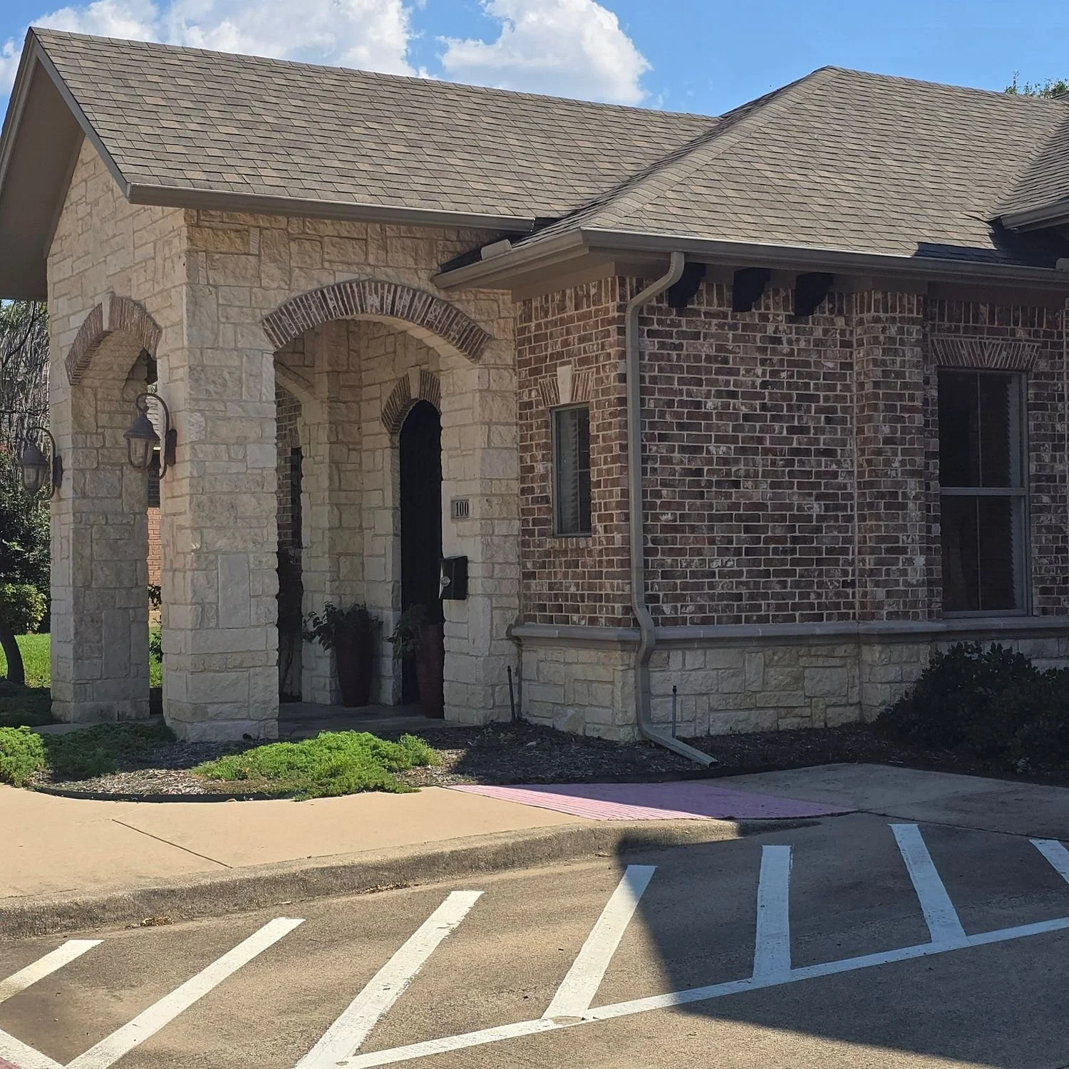 The image shows the exterior of a house with a combination of brick and stone facade, a gabled roof, and a covered entryway supported by stone columns. There are two large planters near the front door, some landscaping at the base of the house, and adjacent parking spaces with white painted lines on the asphalt.