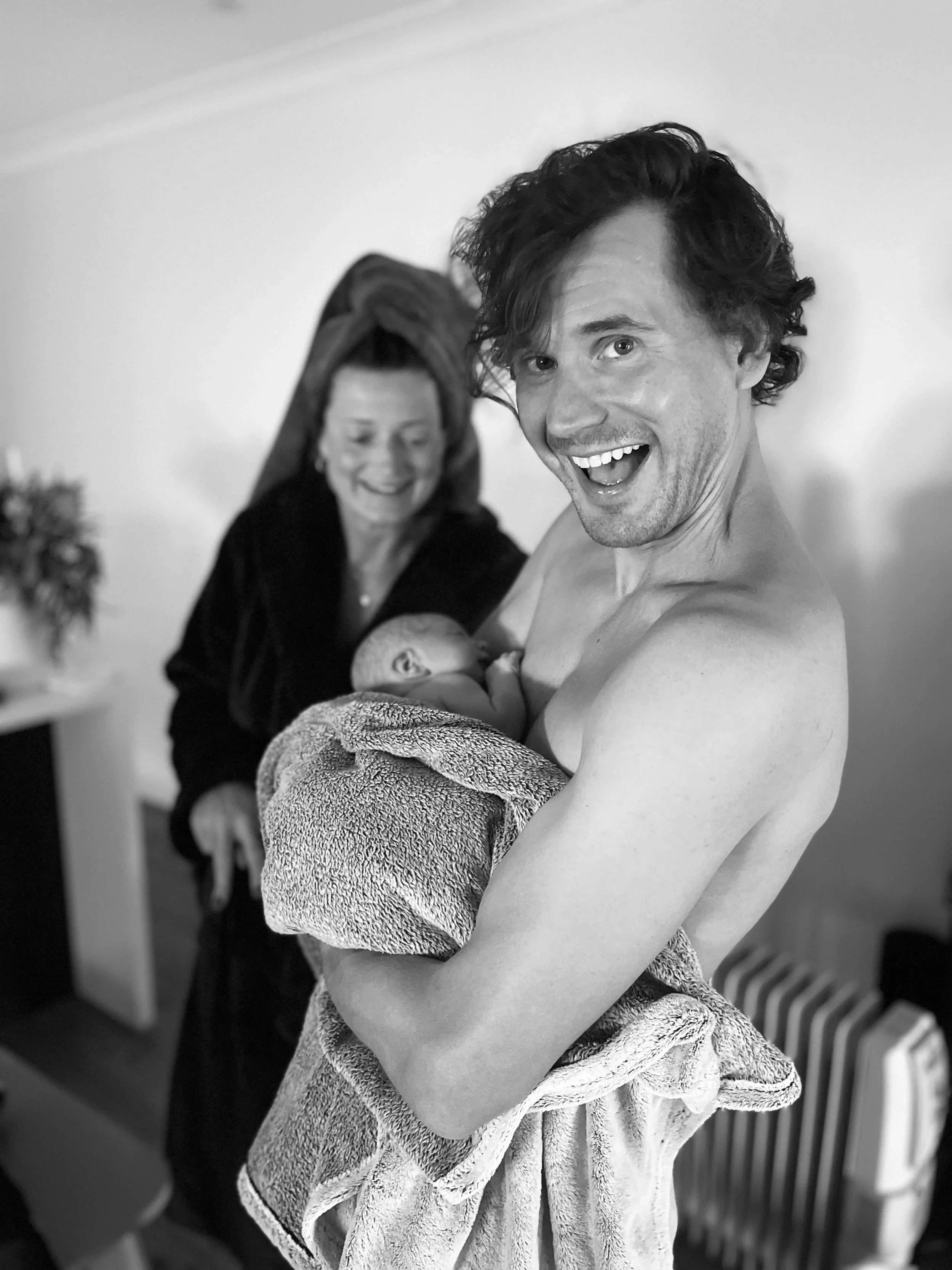 A joyful man with curly hair holding a newborn wrapped in a towel, with a smiling woman in the background in a home setting.